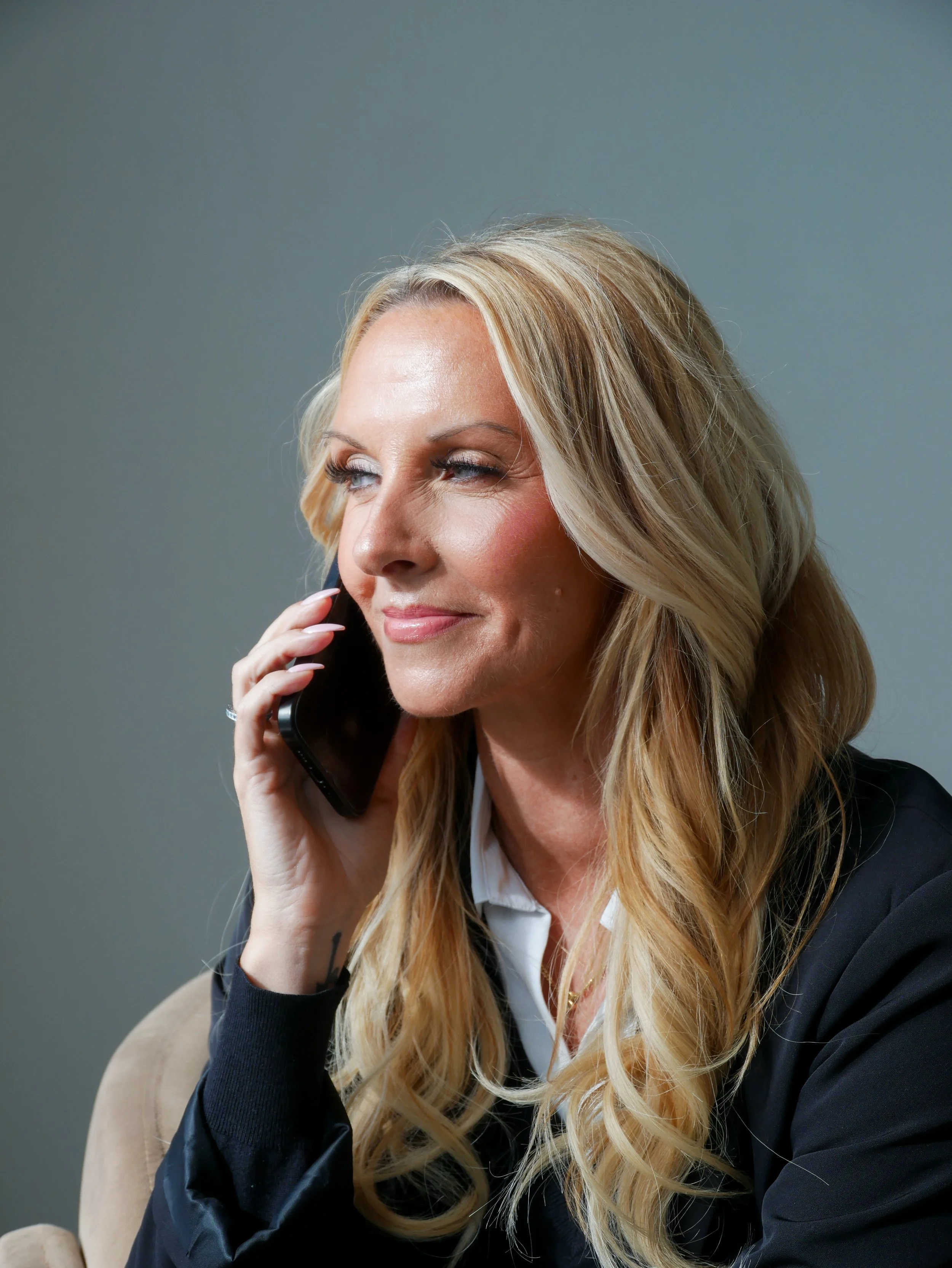 A woman with long blonde hair, wearing a black blazer, is talking on a cellphone. She has a slight smile and appears to be in a professional setting.