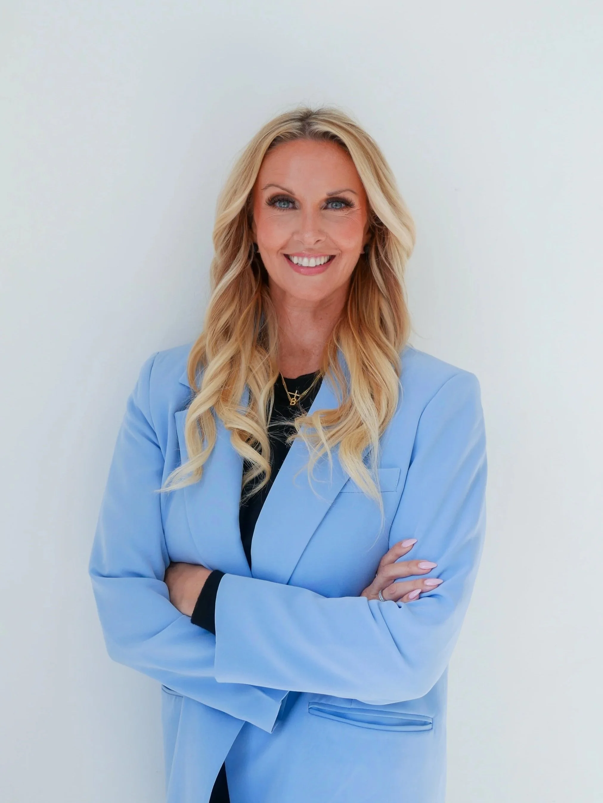 A smiling woman with long blonde wavy hair, wearing a light blue blazer over a black top, standing against a plain white background.