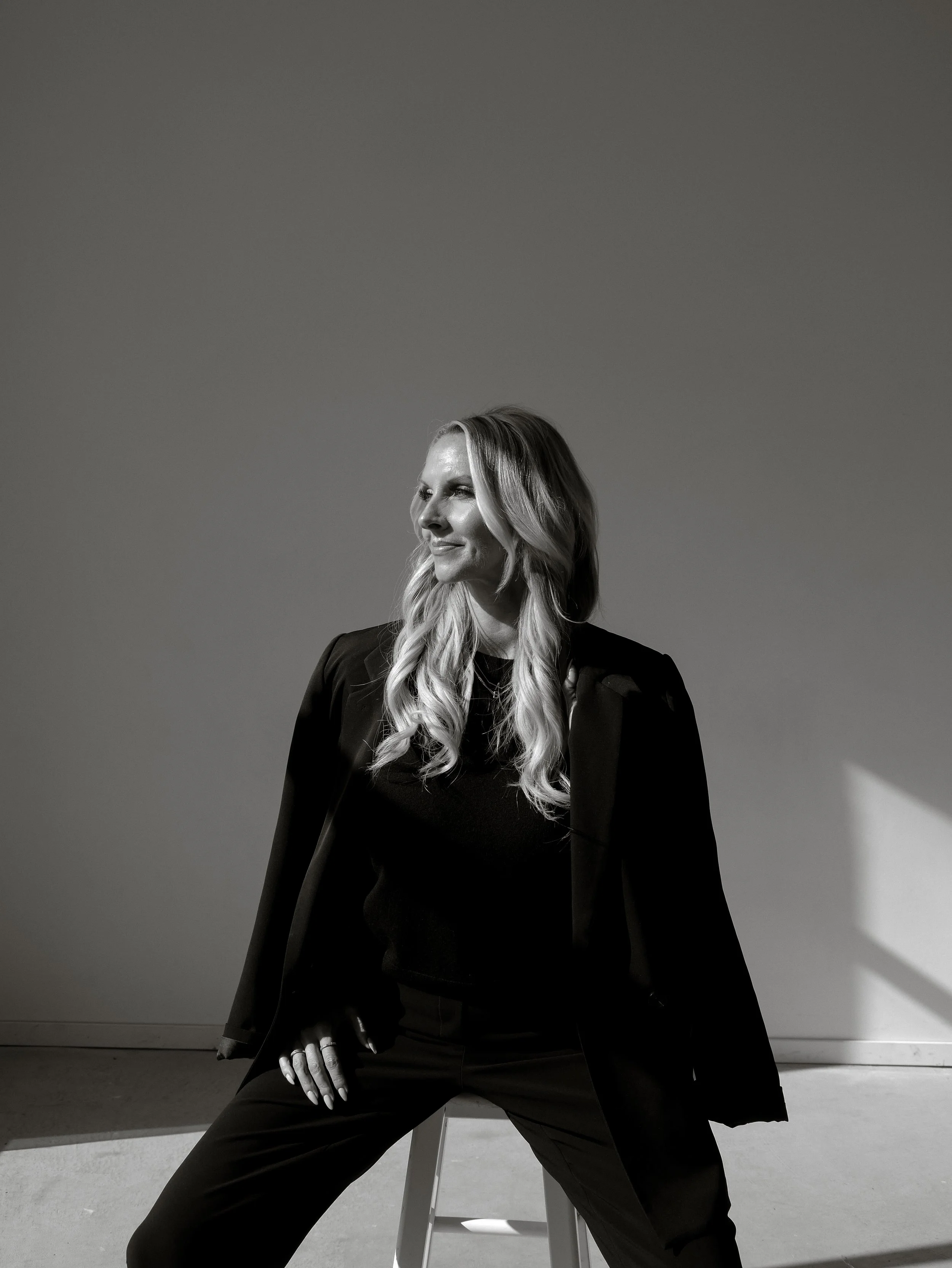 Black and white photo of a woman with long wavy hair wearing a blazer, sitting on a stool, looking to her left with a confident expression against a plain background.