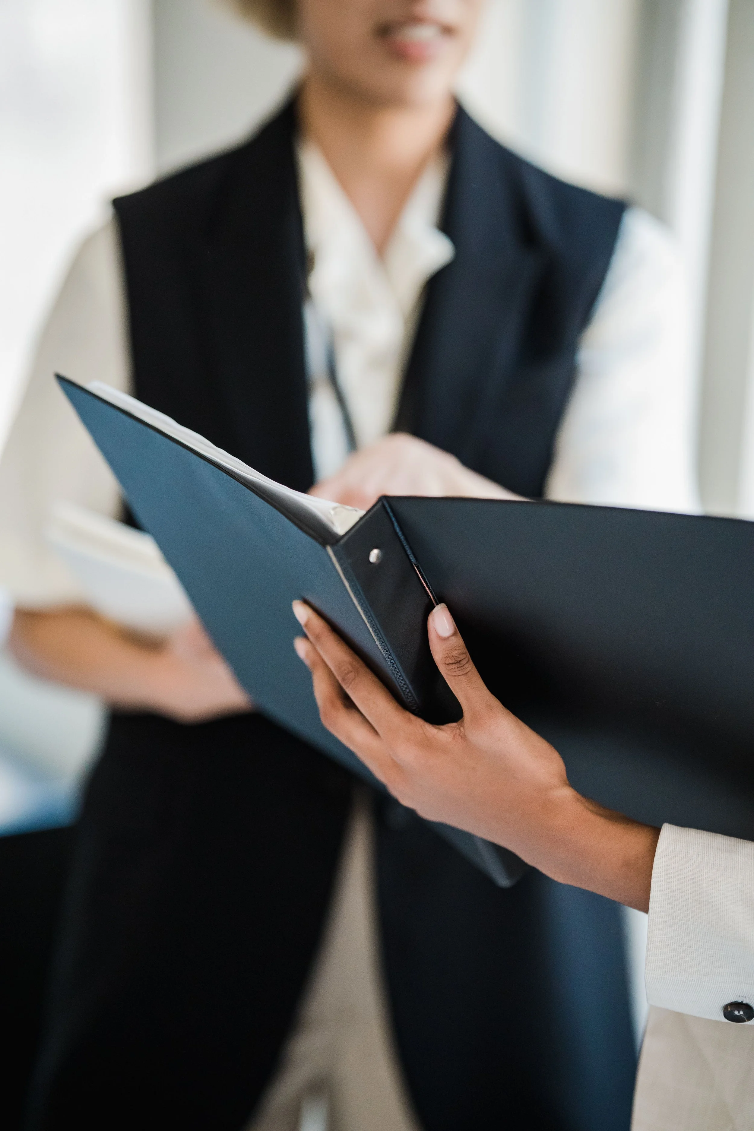 Two people are exchanging documents in an office setting. One person is wearing a white shirt and a black vest, while the other is wearing a white blazer with black buttons. The focus is on their hands and the documents they are holding.