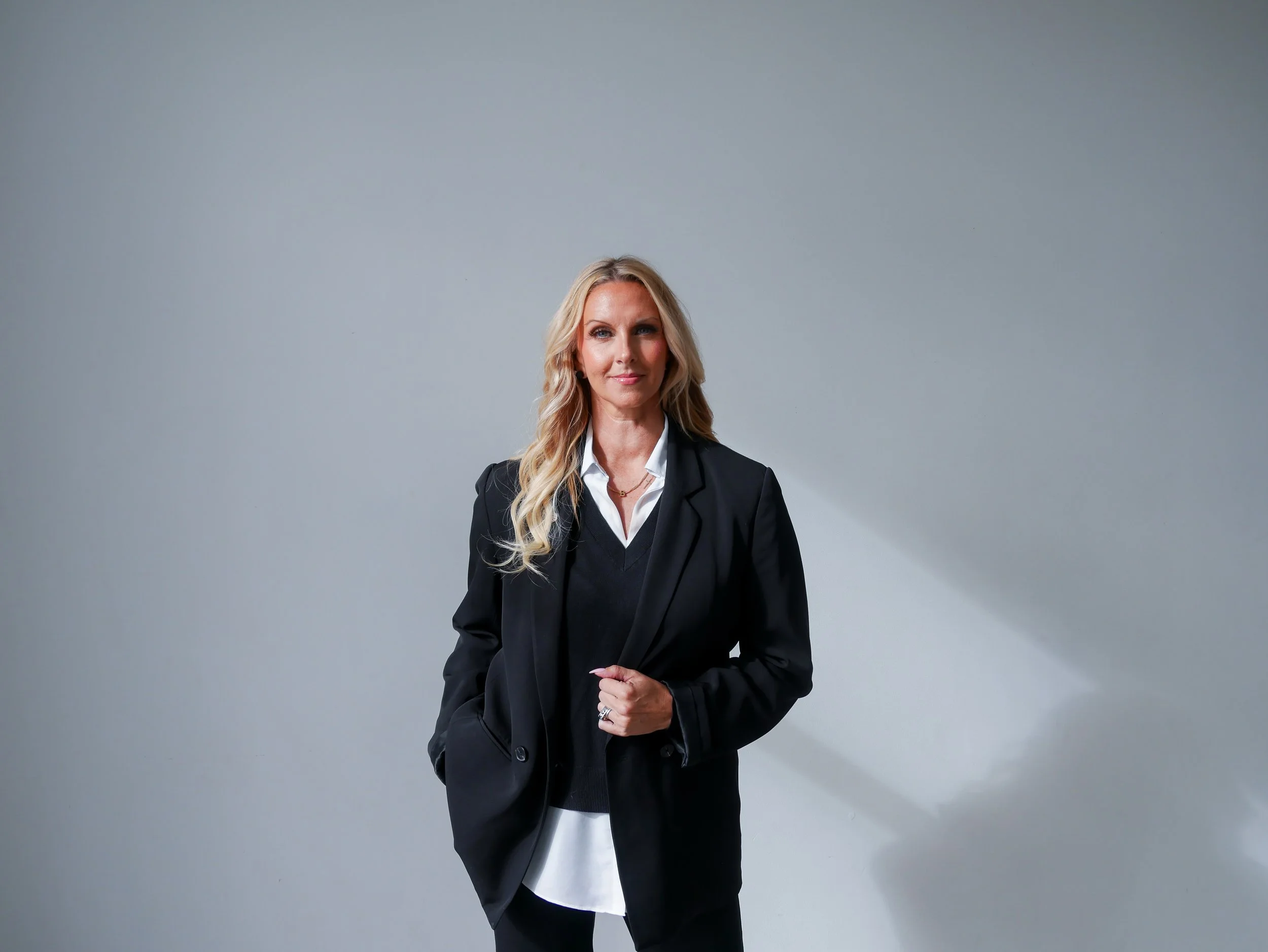 A woman with long blond hair wearing a black blazer over a white shirt standing against a plain gray background.