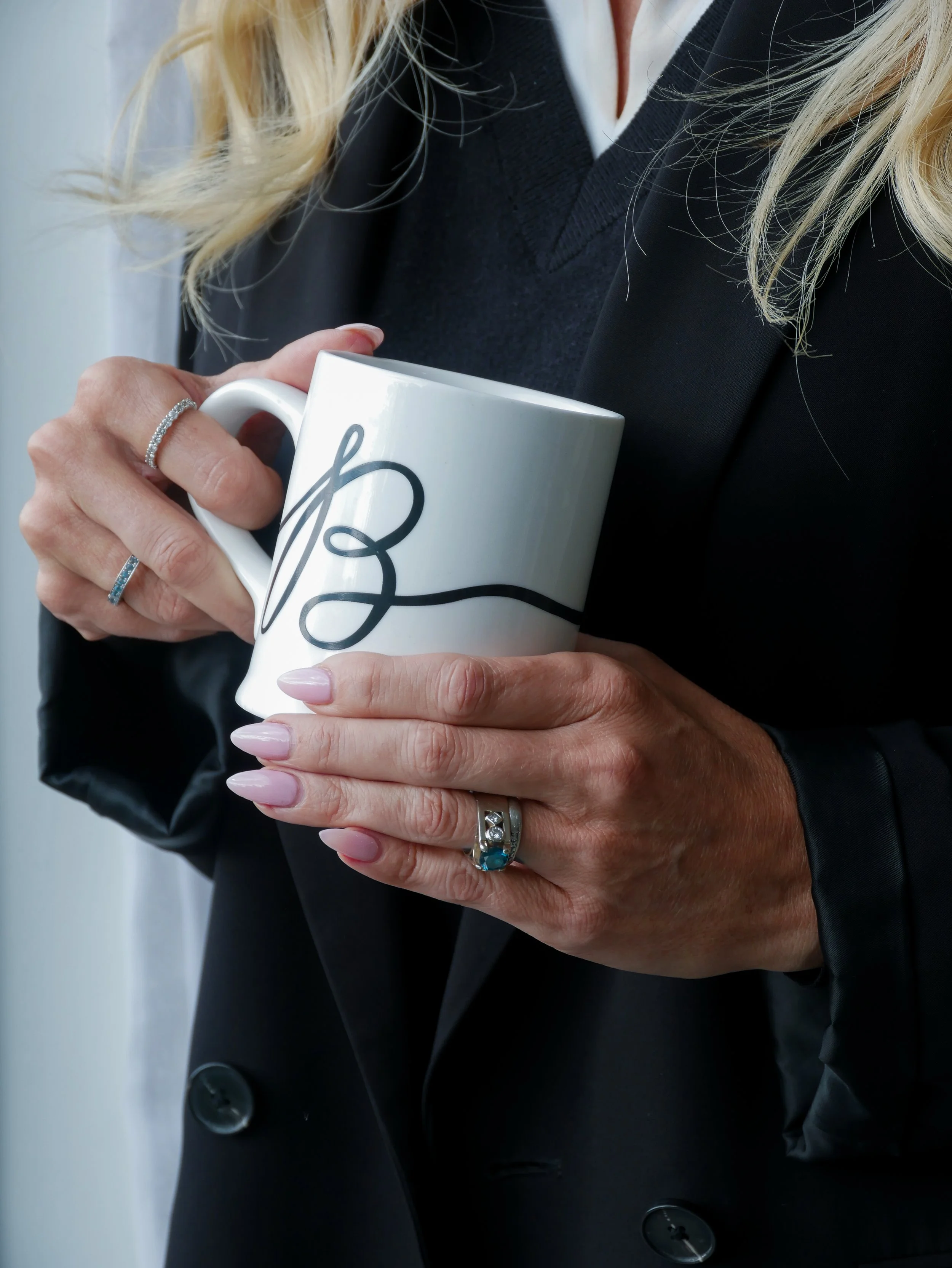 Close-up of a person with blonde hair holding a white mug with black script, wearing a black blazer with large buttons, and two rings on their left hand.