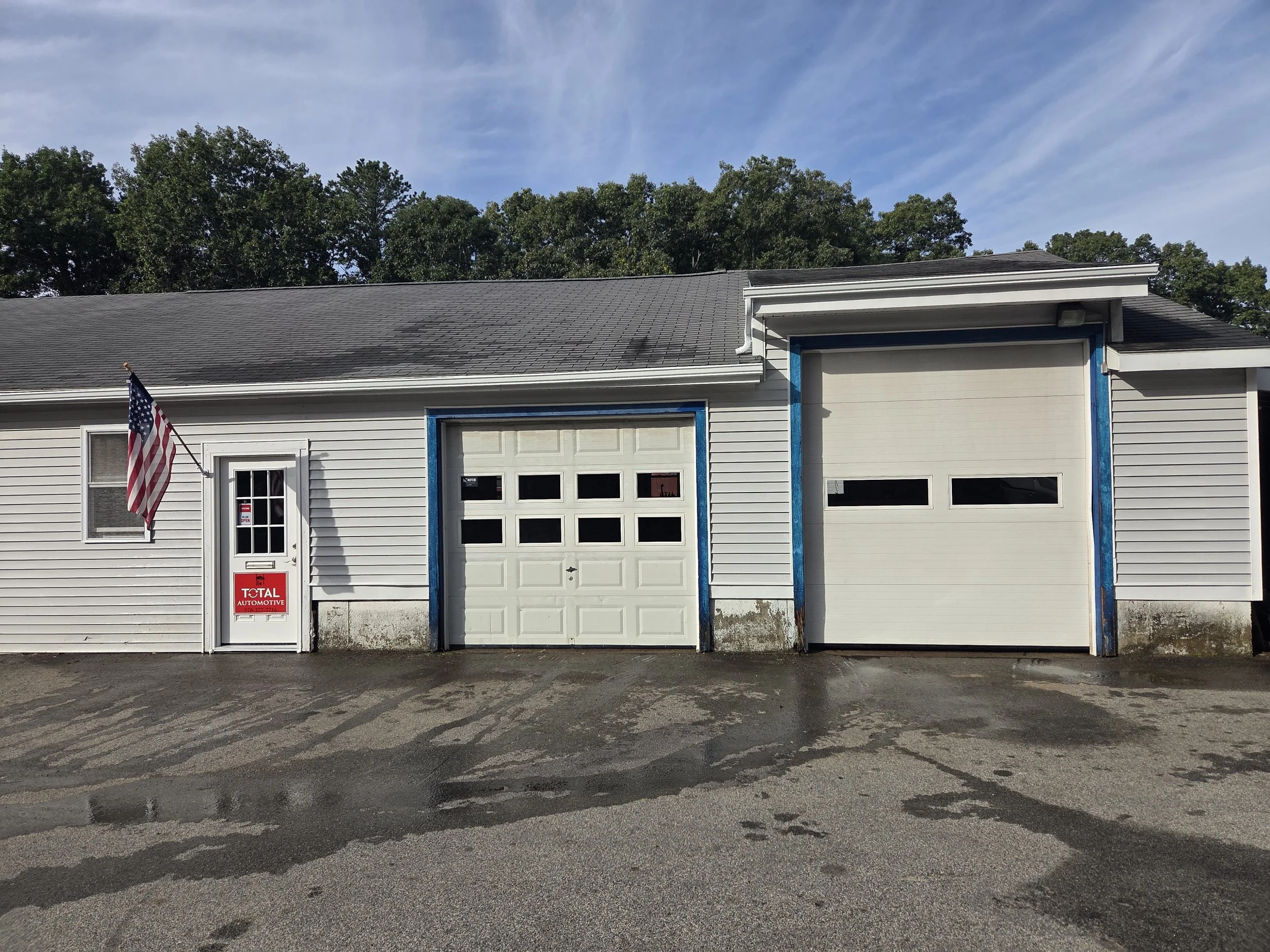 A white garage building with two garage doors and a small door with a window, blue trim around the doors, a red 'Total Automotive' sign, an American flag flag on a pole, a gray paved parking lot, and trees with green leaves under a partly cloudy sky.