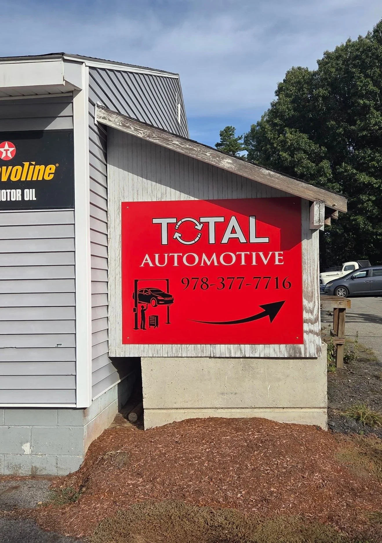 A red sign on a wood wall for Total Automotive with a phone number and a graphic of a car lift and mechanic underneath, located outside a building with a grey siding wall and a tree in the background.