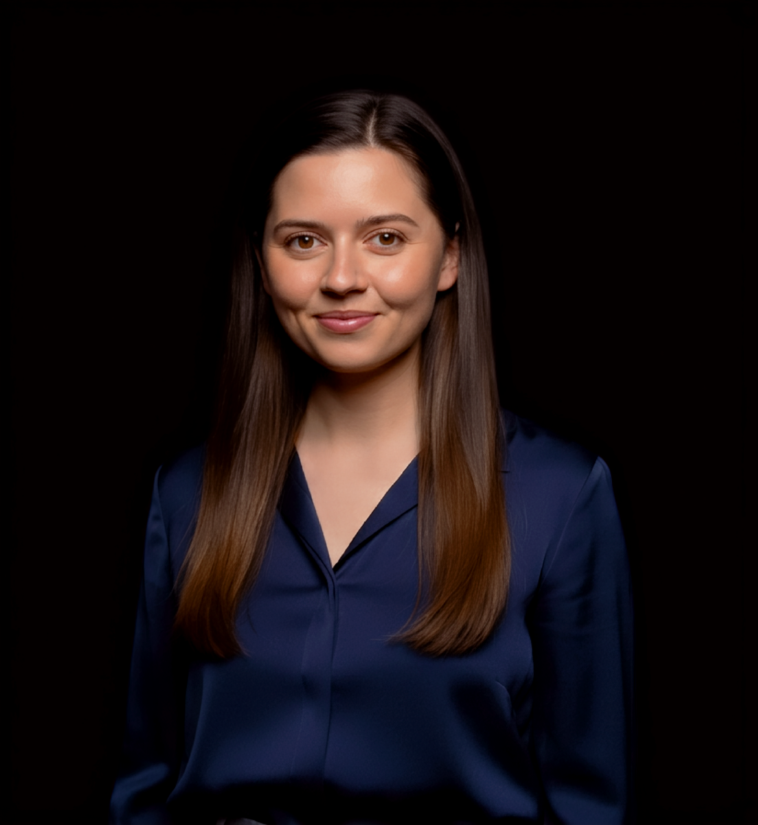 Portrait of a young woman with straight brown hair, brown eyes, and light skin, wearing a dark navy shirt, smiling softly against a black background.