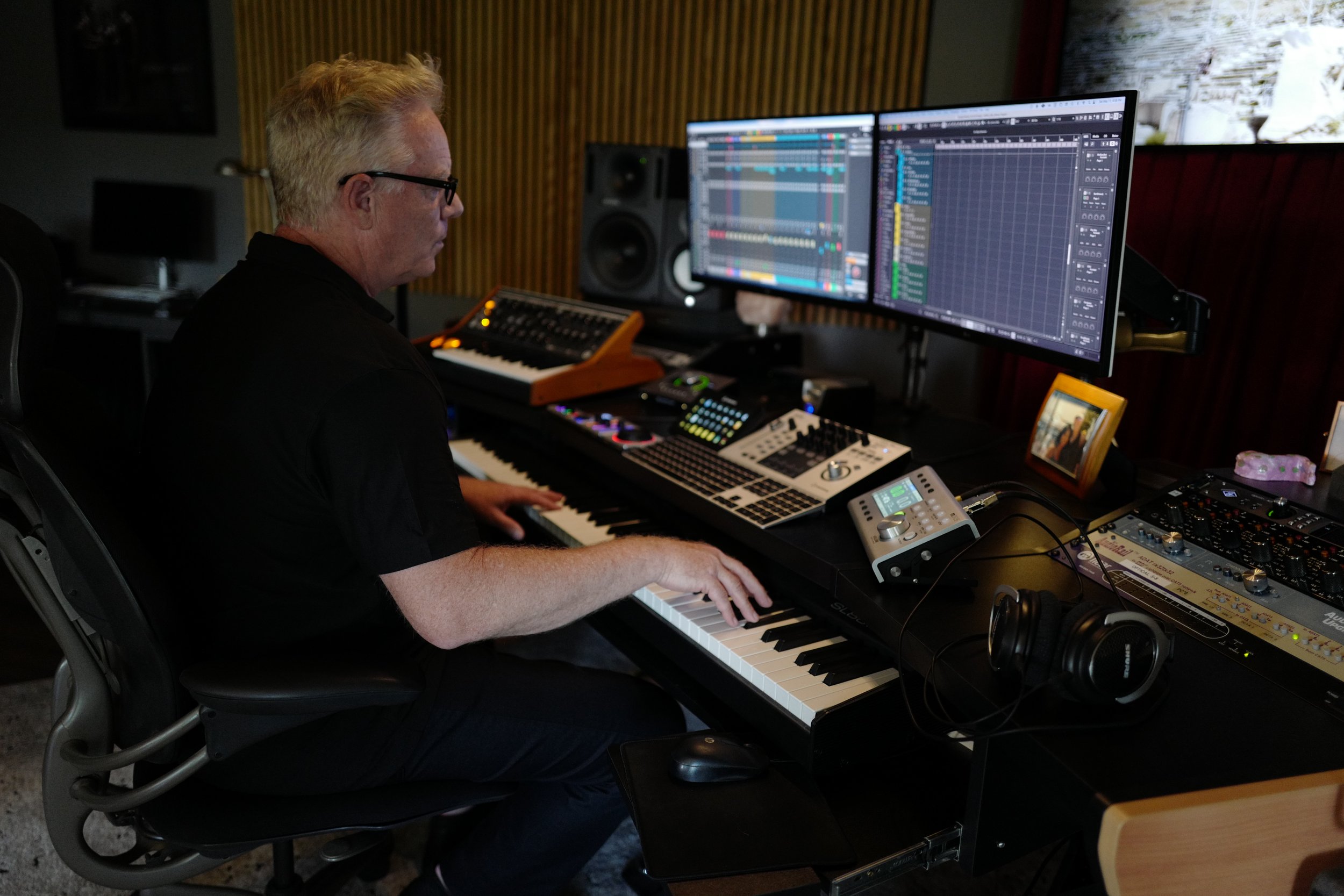 A person with glasses playing a keyboard surrounded by music production equipment, including dual computer monitors displaying music software, an audio interface, and a mixing console, in a recording studio.