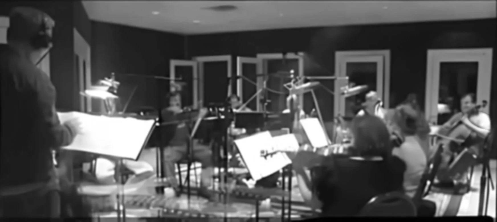 A black and white photo of a group of people in a recording studio, some seated and others standing, with music stands and microphones.