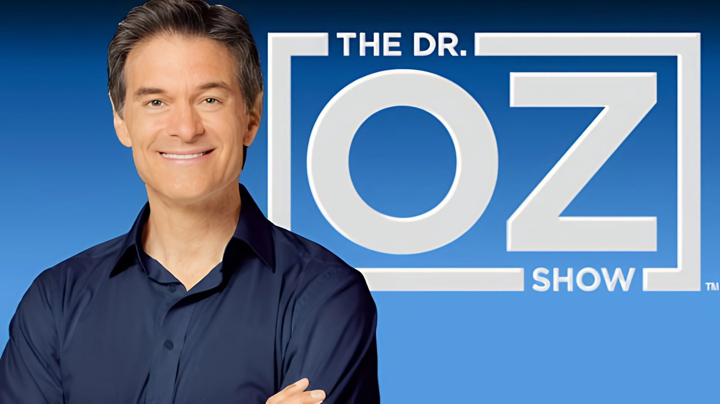 A smiling man in a dark blue shirt standing in front of a blue background with a sign that reads 'The Dr. Oz Show' in white letters.