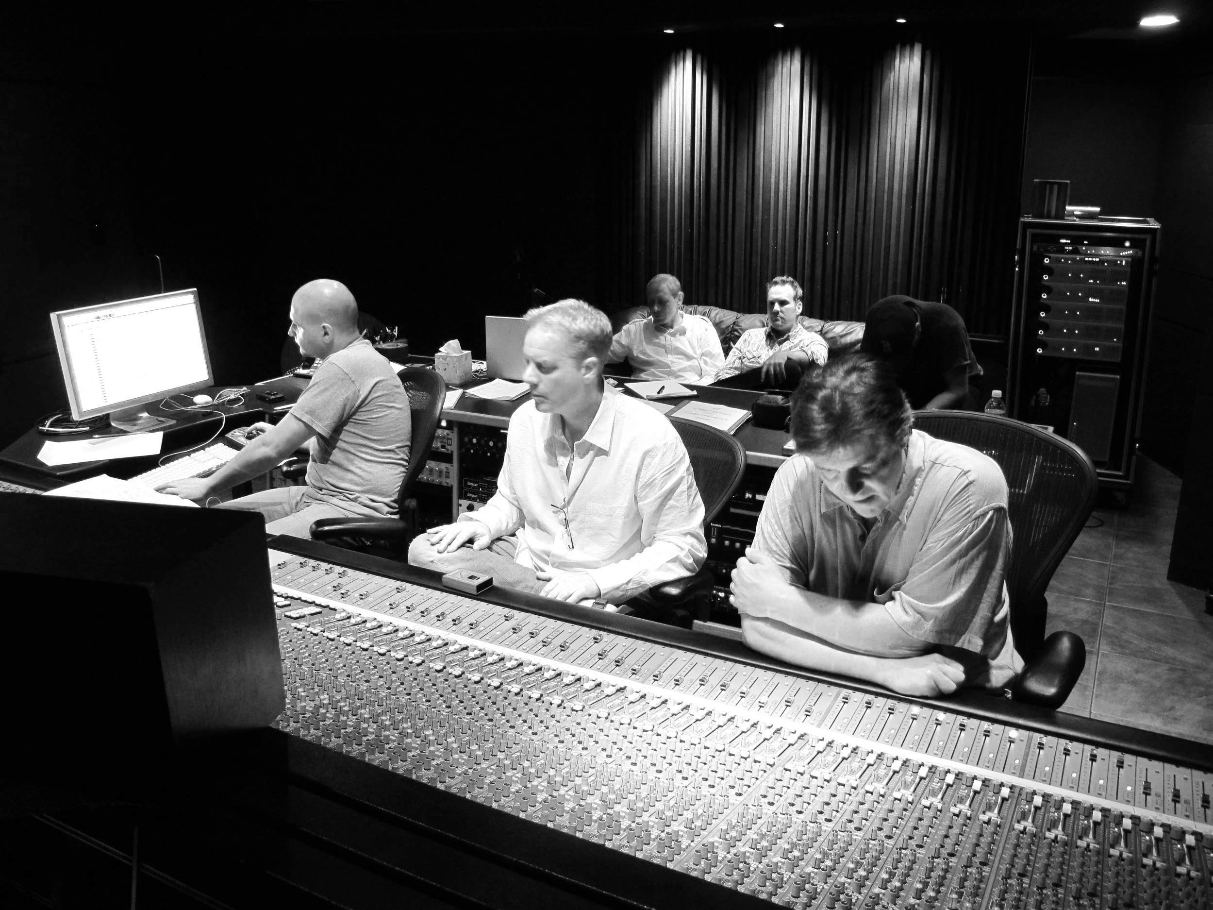 Black and white photo of a group of people working in a recording studio. They are seated at a mixing console and computers, with some of them looking at their screens and others engaged in conversation.