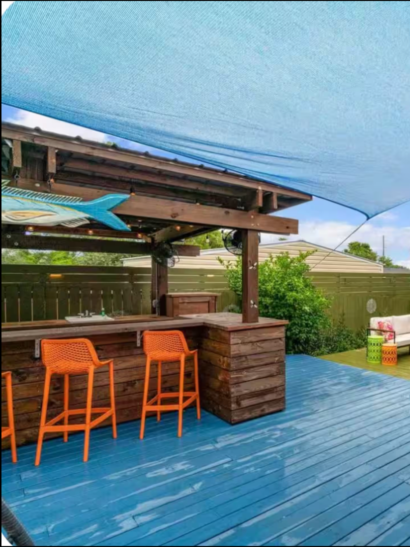 Outdoor patio with a blue wooden deck, a wooden bar with two orange chairs, a blue fabric shade, and green foliage in the background.