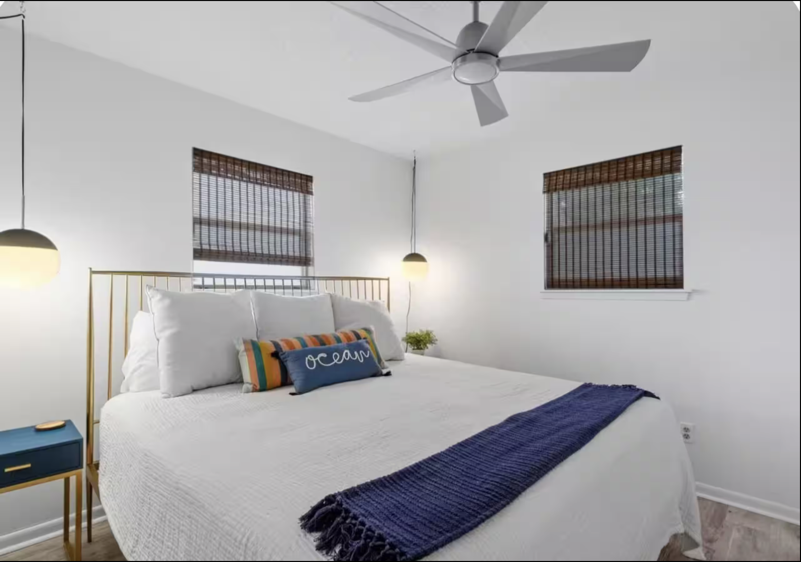 A bedroom with a white bed, navy blue blanket, and pillows, two windows with wooden blinds, a ceiling fan, a bedside lamp, and a small blue bedside table with a plant.