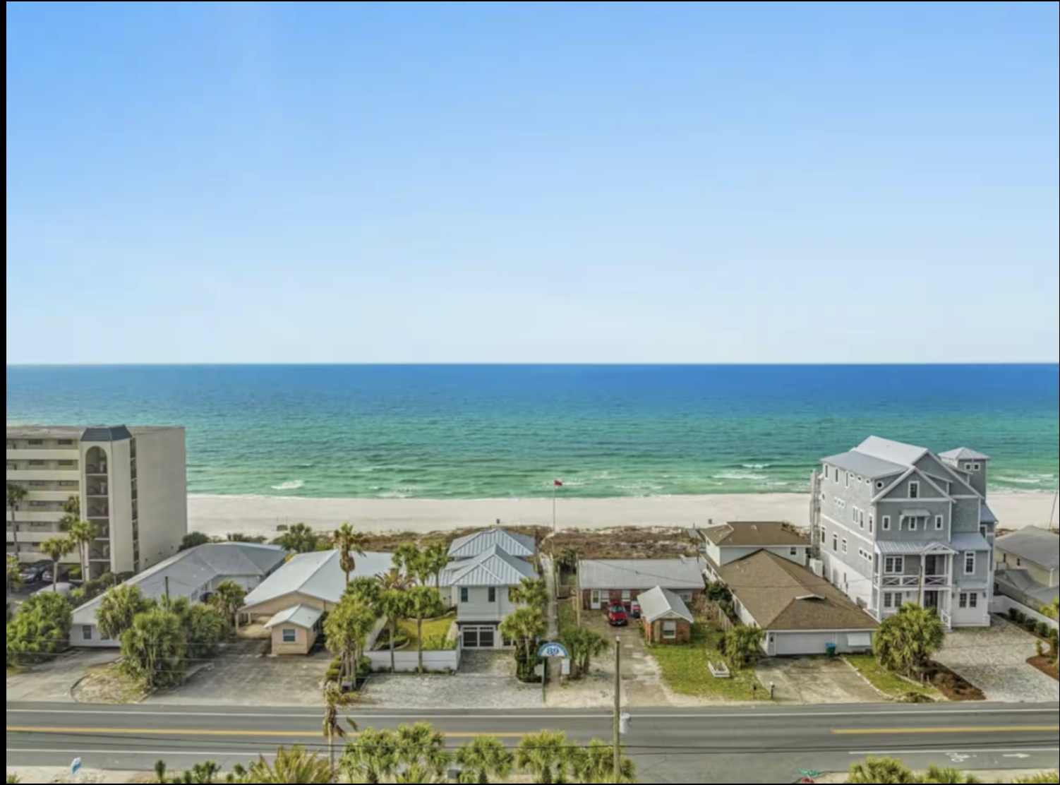A coastal scene with a view of the ocean, sandy beach, and residential houses and buildings along the shoreline with a clear blue sky.