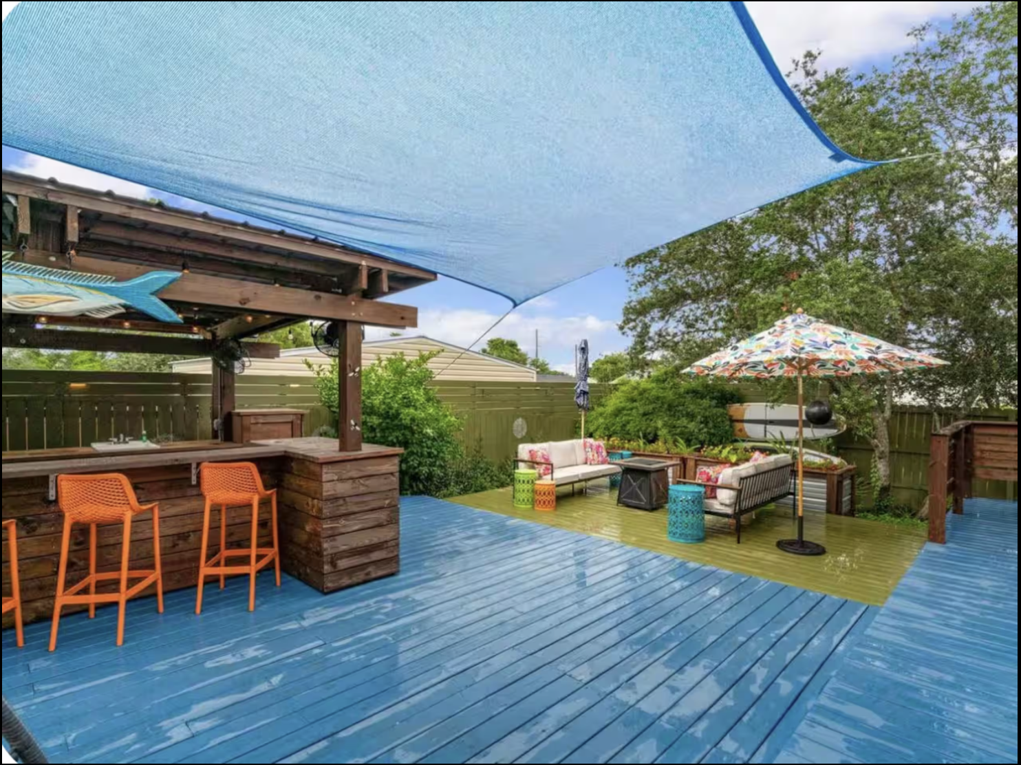An outdoor backyard patio area with a blue wooden deck, a shaded seating area with cushioned furniture and colorful pillows, an umbrella with a floral pattern, and a small bar area with orange chairs. There is greenery and trees in the background.