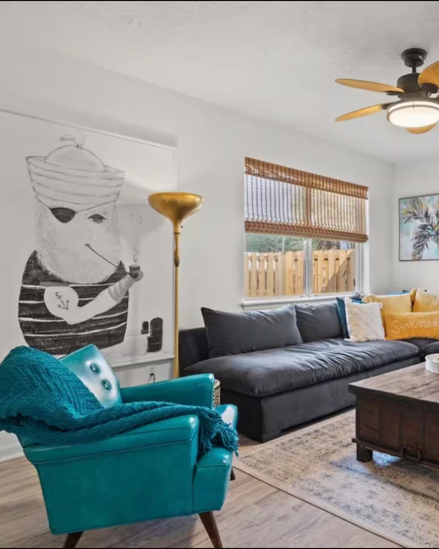 Living room with a black sofa, a turquoise armchair with a blanket, a wooden coffee table, a gold floor lamp, a ceiling fan, a window with wooden blinds, and a framed artwork of a palm tree on the wall. There's a large black-and-white mural of a person with a turban, sunglasses, and smoking a pipe.