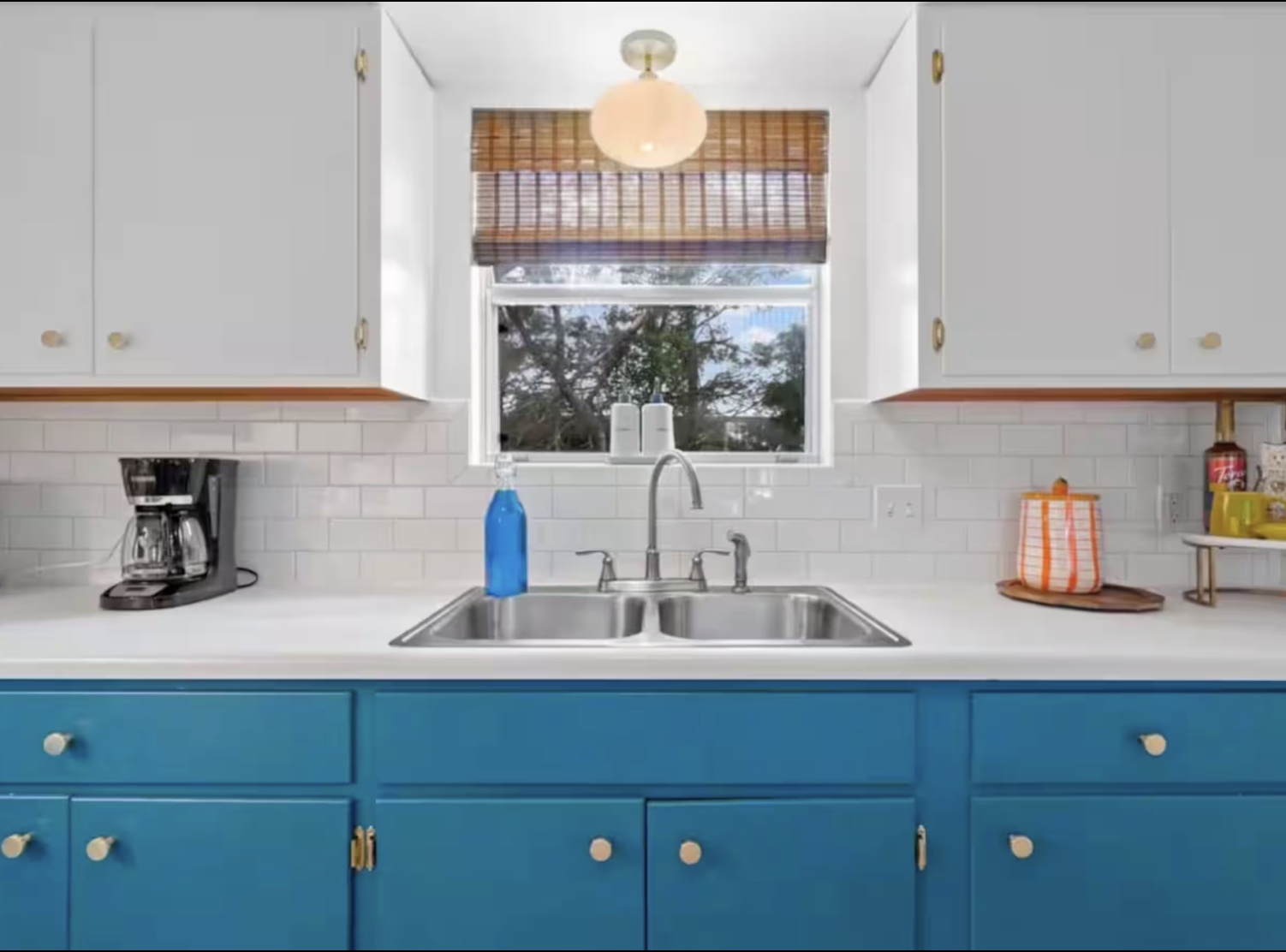 Kitchen sink with a blue dish soap bottle, a window with a bamboo blind, white cabinets, and a blue lower cabinet. Items include a coffee maker and a paper bag on the countertop.