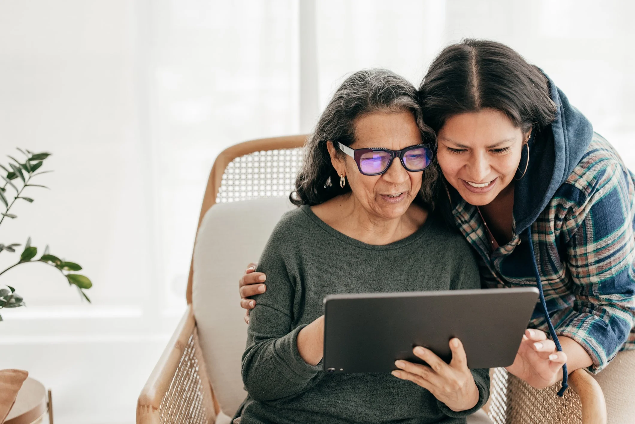 An elderly woman with gray hair and glasses sitting on a chair, smiling and looking at a tablet held by a younger woman with dark hair. The younger woman is leaning in, smiling, and wearing a plaid shirt. They are in a bright room with a white wall and a plant on the left.