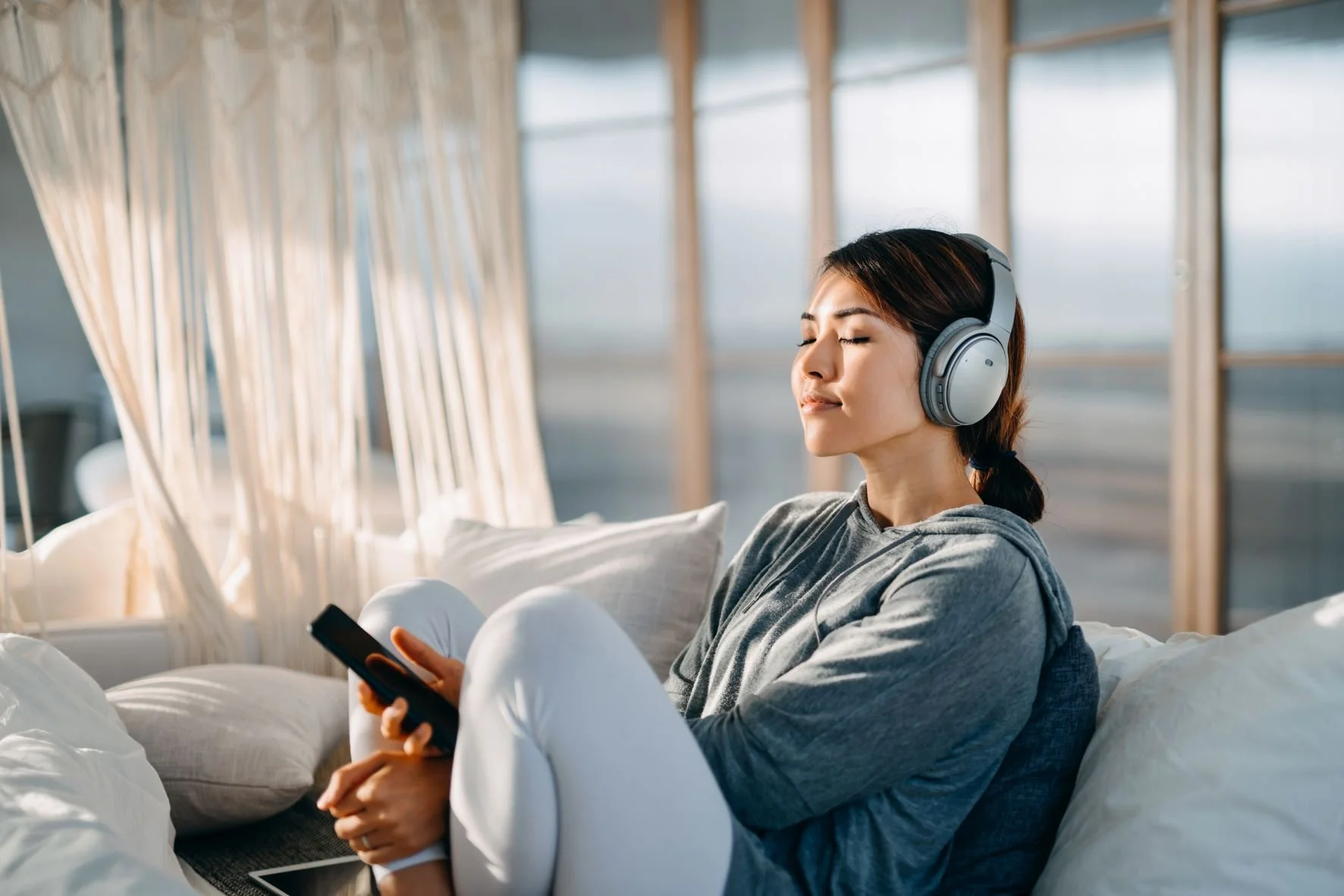 Young woman with dark hair wearing headphones and a gray hoodie, sitting on a bed/or lounge area in a modern, open space, holding a smartphone with a relaxed expression.