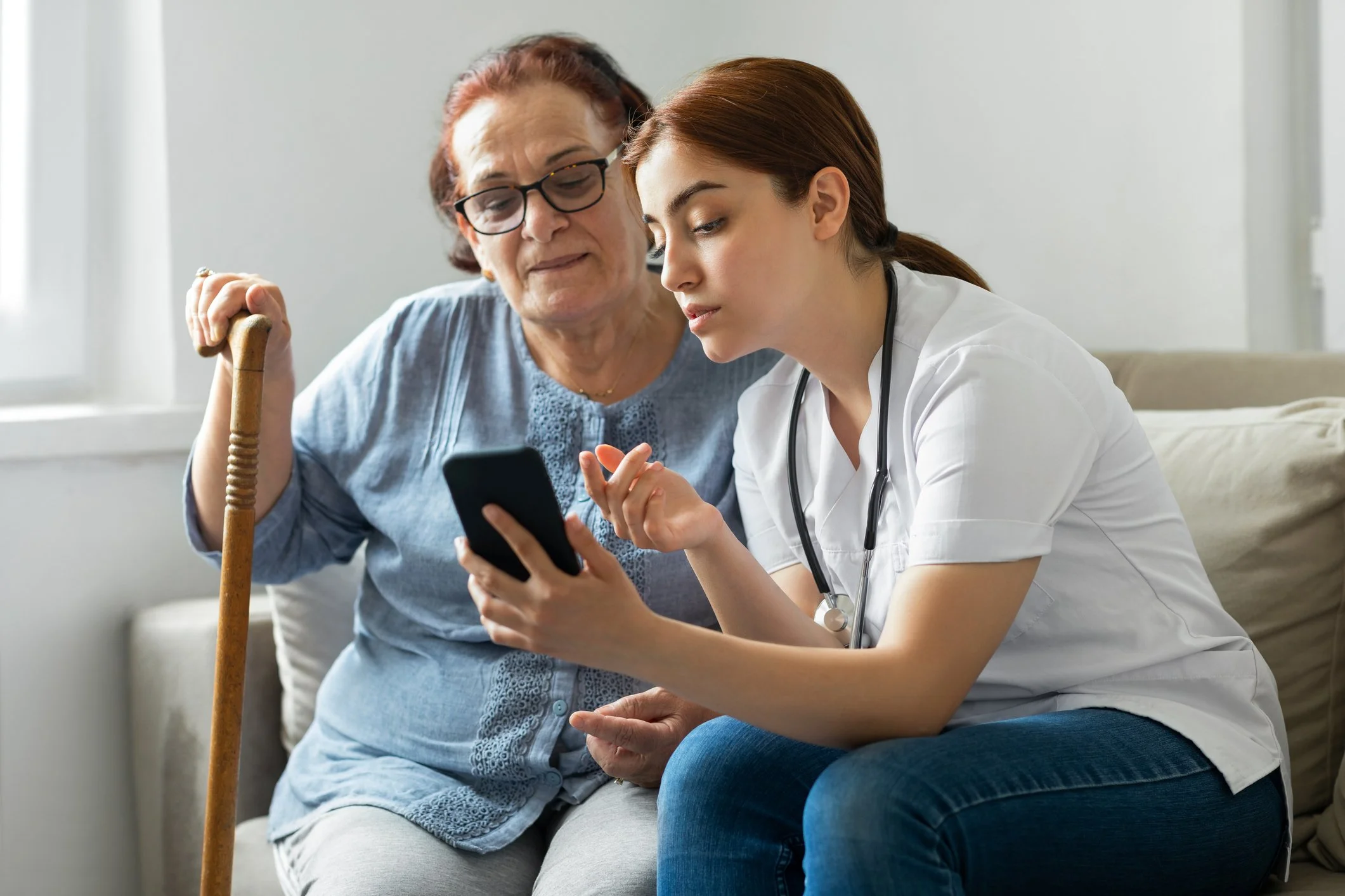 A healthcare professional, likely a nurse or doctor, wearing a white coat and stethoscope, assisting an elderly woman with a cane by showing her something on a smartphone.