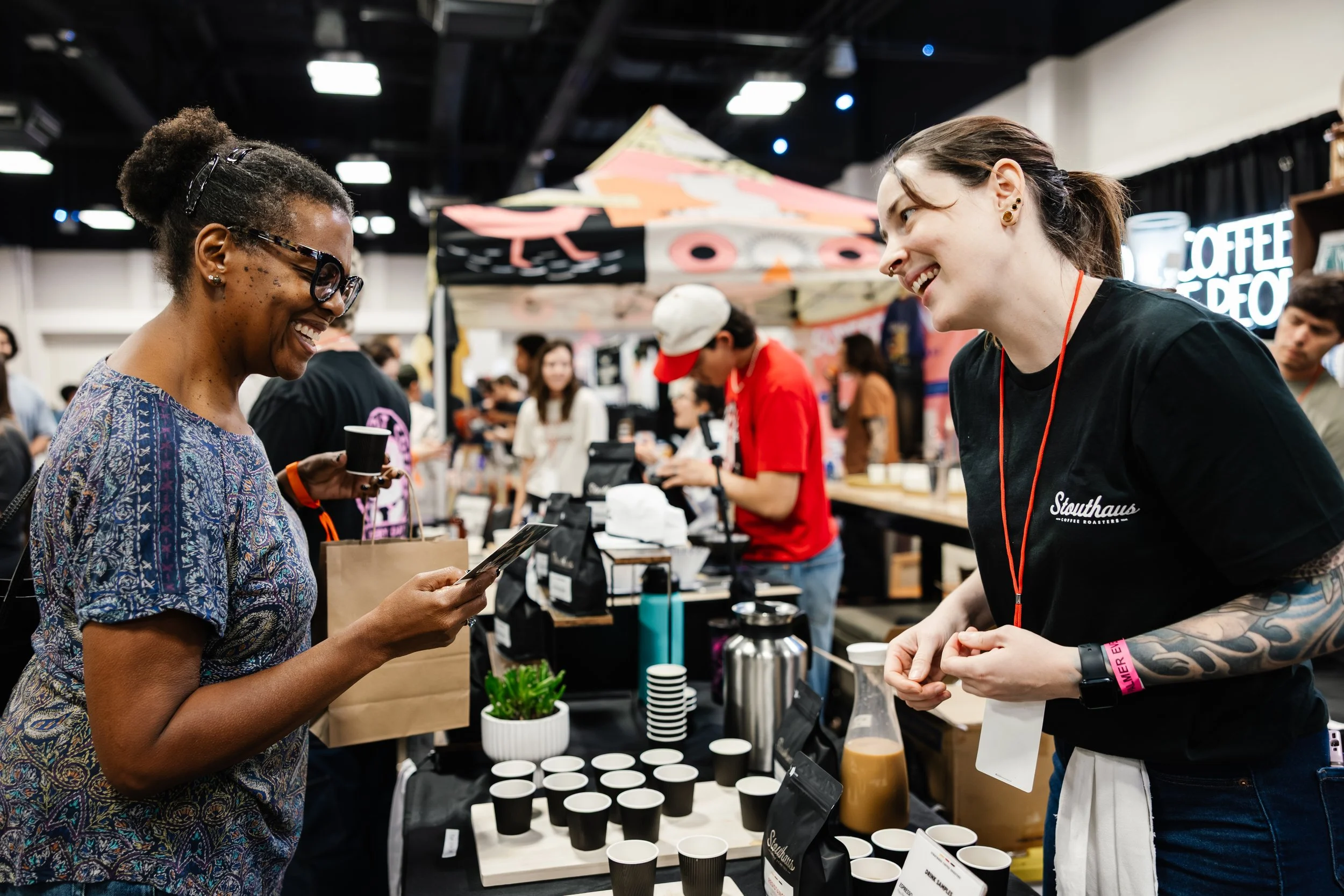 Coffee vendor smiling and talking with an attendee while serving tasting samples at a coffee festival booth.