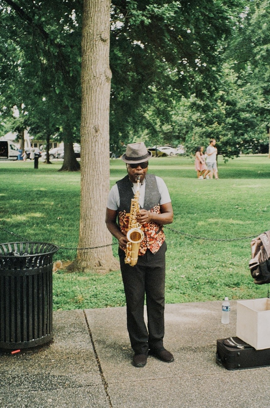 Street saxophonist wearing a fedora and vest performs in a green park.