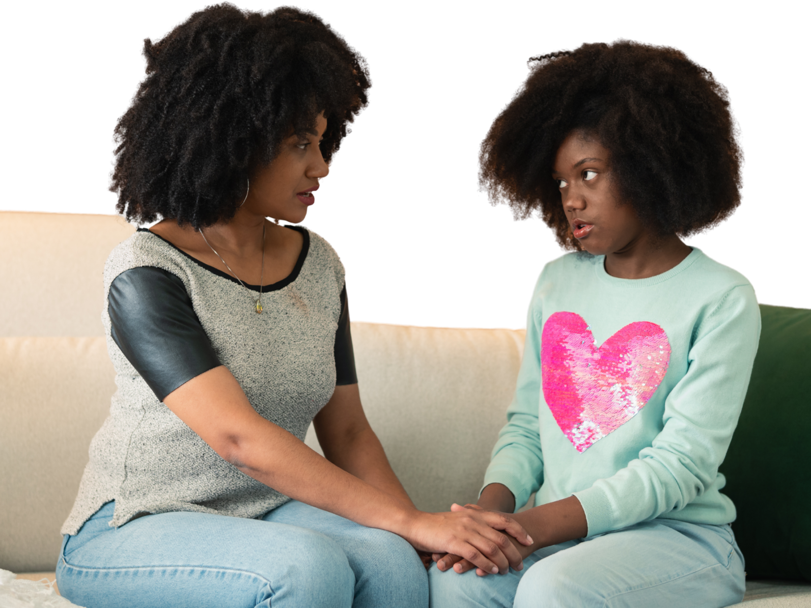 An adult woman and a young girl sit closely on a beige couch, holding hands and looking at each other.