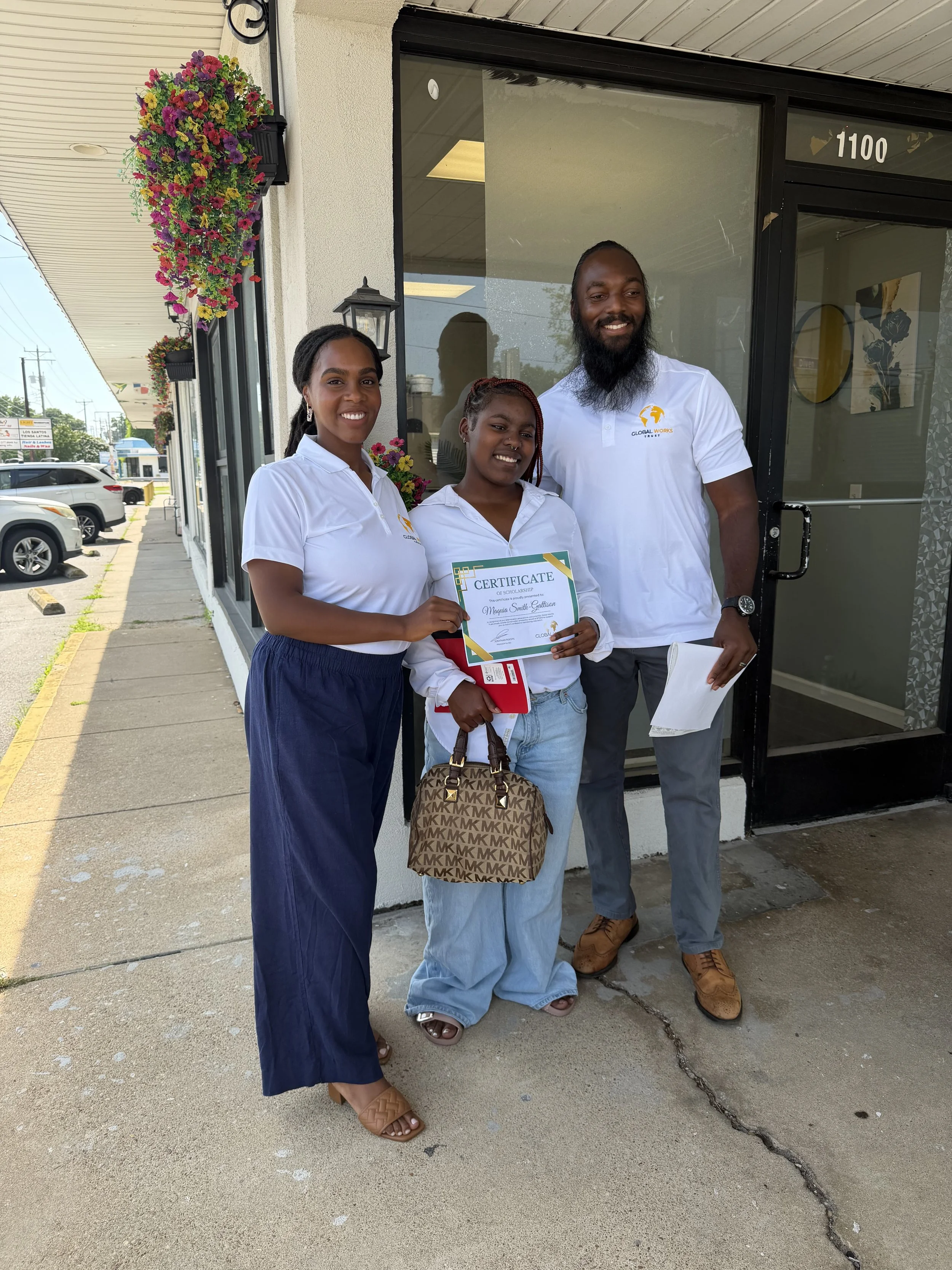 Jonathan and Camarie, founders of CFFYD are presenting a $17,500 scholarship. Camarie is holding a certificate of scholarship. They are all wearing white shirts with a logo. The building has hanging flower baskets and large window.