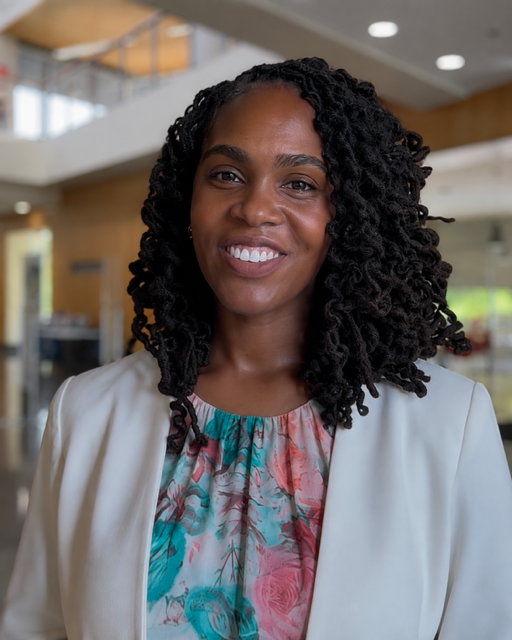 Smiling woman with curly hair wearing a floral blouse and white blazer indoors.