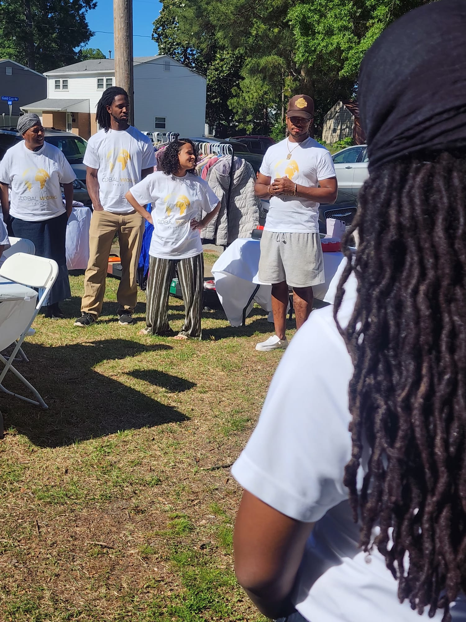 People gathered outdoors at a community event, some wearing shirt with a yellow globe logo that says 'Global Works'. They are standing near tables and clothing racks on a sunny day.