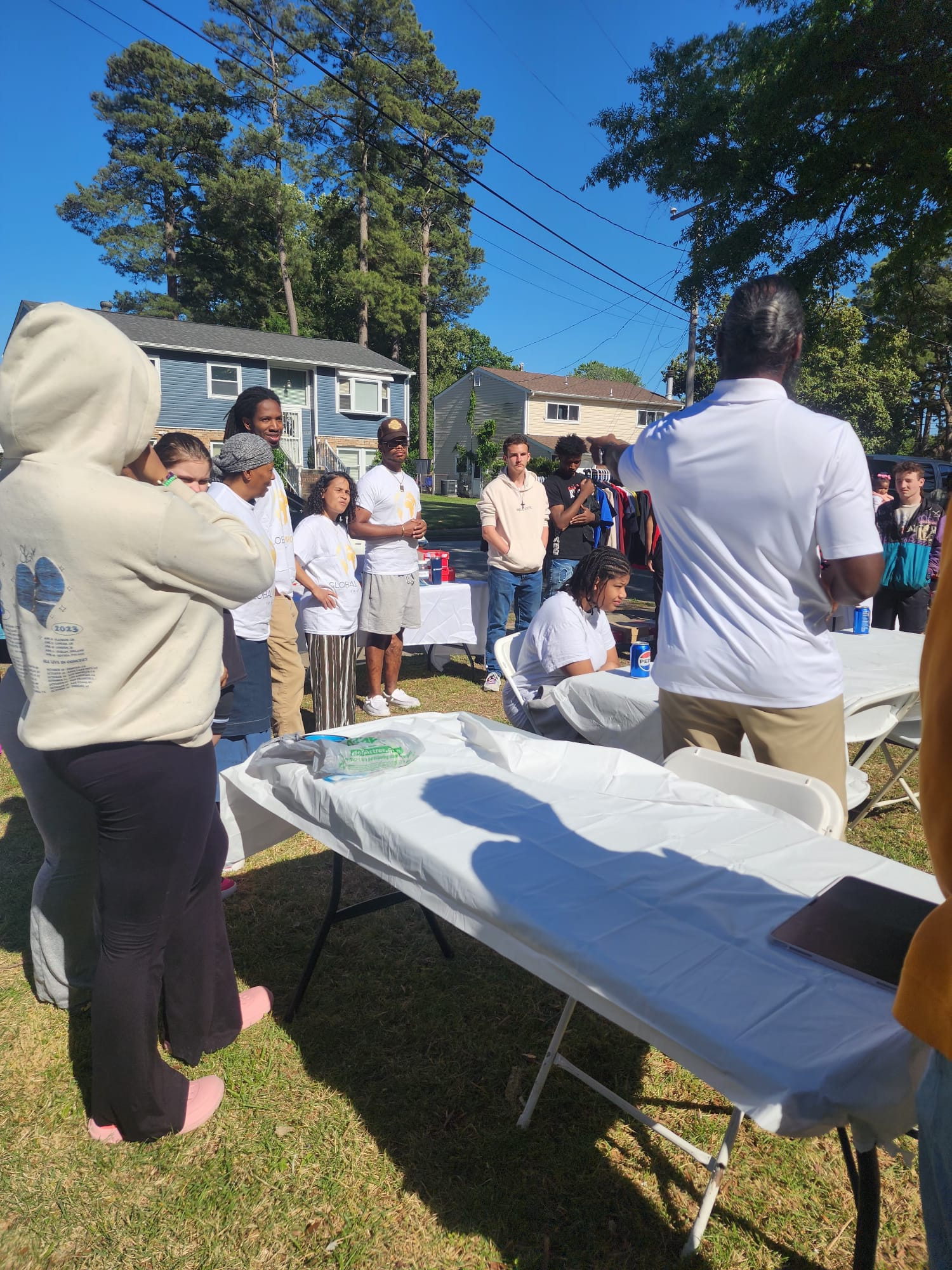 A group of people gathered outdoors around folding tables, some standing and some sitting, with a blue house, green trees, and a clear sky in the background. It appears to be a community event or gathering.