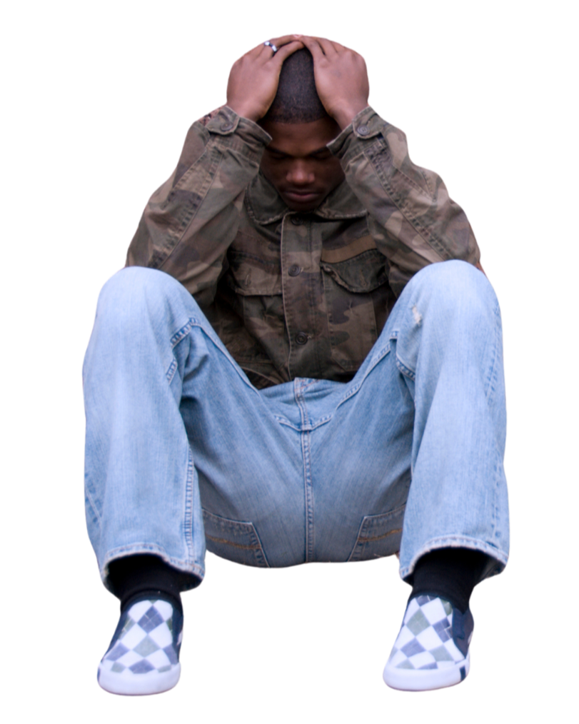 A young man sitting on the ground, holding his head with both hands, wearing a camouflage jacket, light blue jeans, and checkered slip-on shoes.