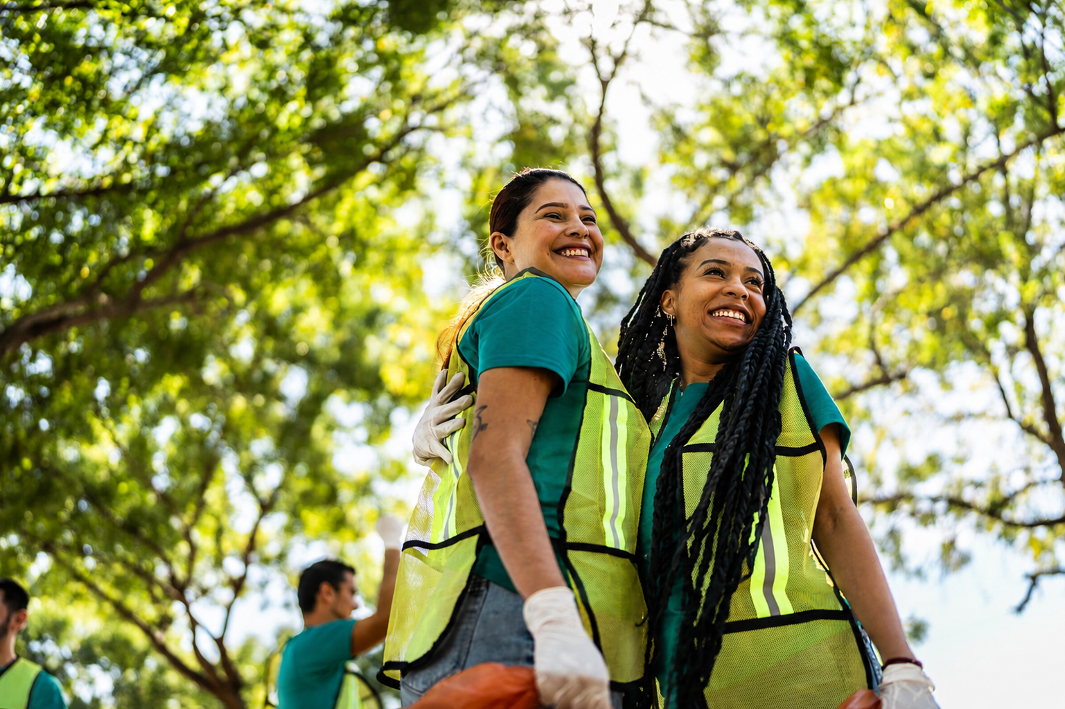 Two women in yellow safety vests smiling outdoors, with trees and blue sky in the background.