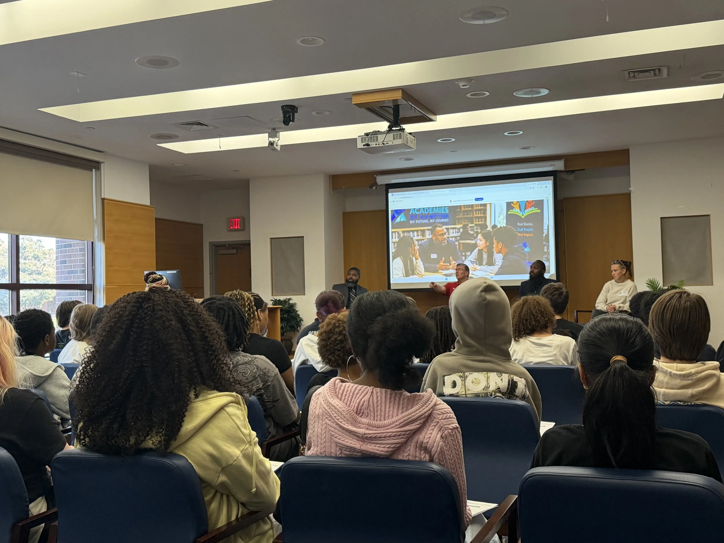 A diverse group of people attending a presentation or panel discussion in a conference room with a large screen displaying a logo and a photo of a group of people in a library setting.