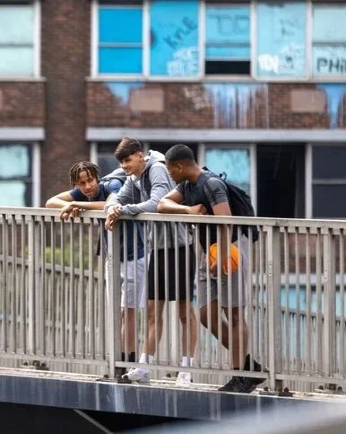 Three young men stand on a bridge, leaning on the railing and looking down at something below, in an urban area with modern brick buildings and large glass windows behind them.