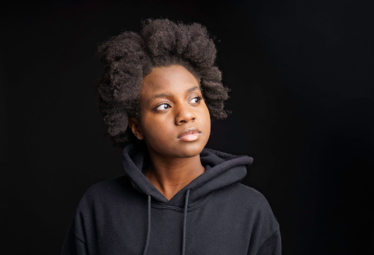 Portrait of a young woman with natural curly hair wearing a black hoodie, looking to her left against a black background.