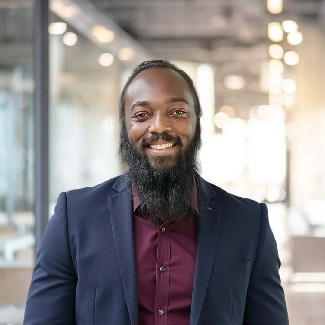 A smiling man with a beard wearing a navy blazer and a maroon shirt standing in a modern office setting with glass walls and bright lighting.