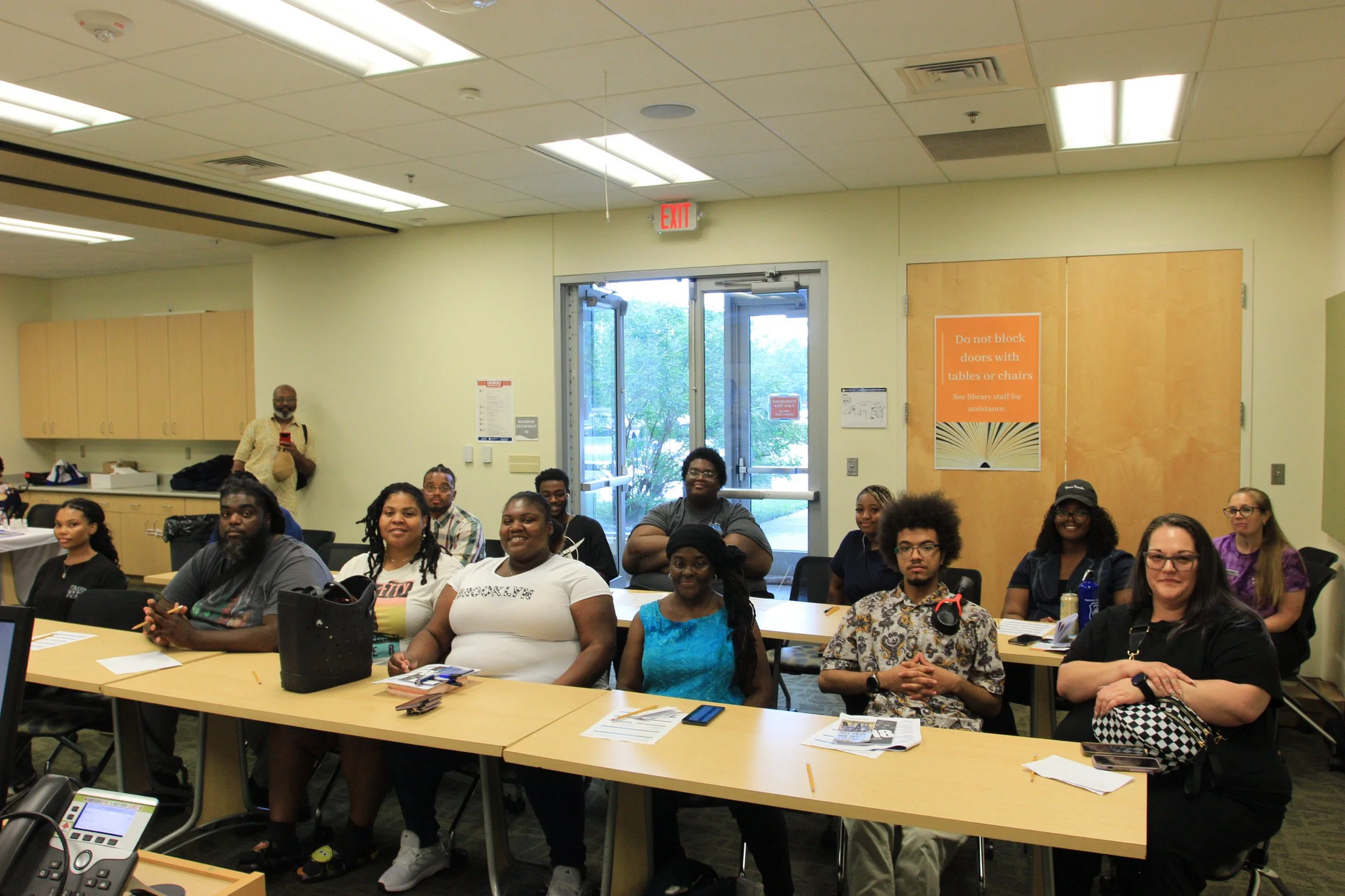 A diverse group of people sitting at tables in a conference room, some smiling at the camera, with a door and exit sign in the background.