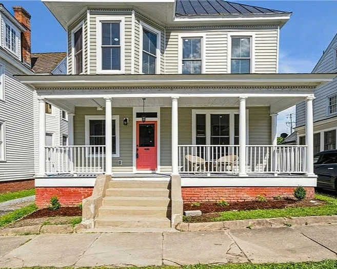 Front view of a two-story house with a porch, white siding, and a red front door. There are steps leading up to the porch and outdoor seating on it, with a small garden bed along the front.