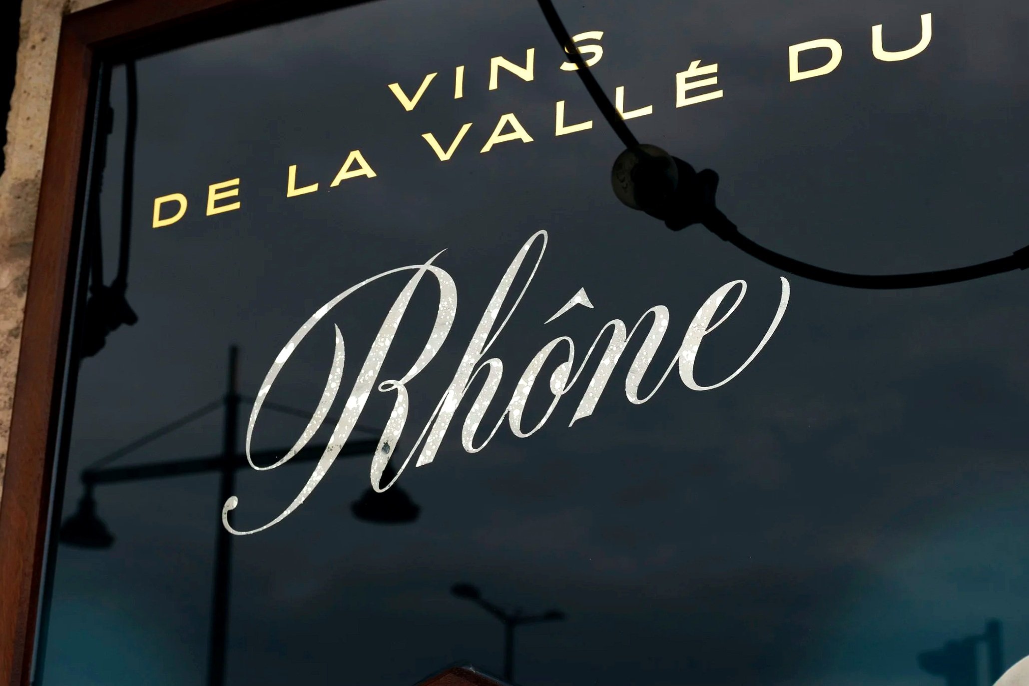 Une enseigne en verre avec des lettres en écriture cursive et moderne indiquant 'VINS DE LA VALLEE DU RHÔNE'. réalisé en dorure à la feuille d'or et feuille d'argent avec des effets de 'gouttes' dans le lettrage.