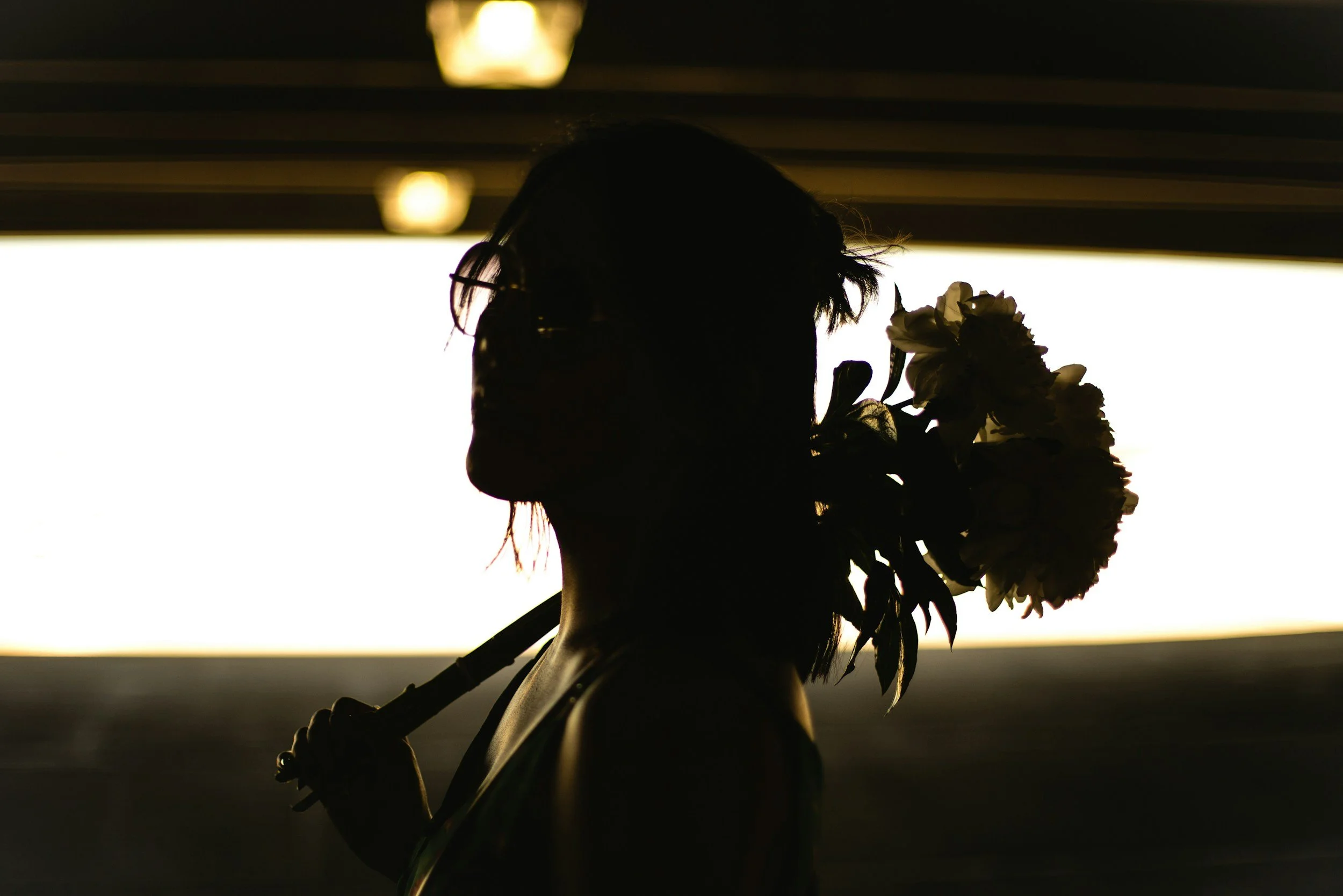 Silhouette of a woman holding flowers, with glasses and short hair, against a backlit background.