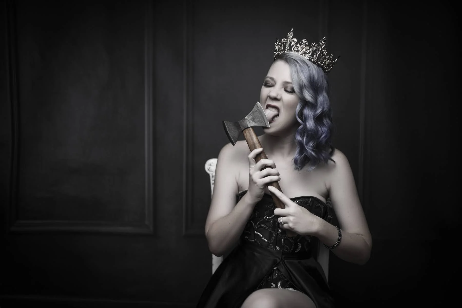 A woman with wavy purple hair wearing a crown and black strapless dress, sitting in front of a dark background, holding an axe near her face, with her tongue out licking the axe.