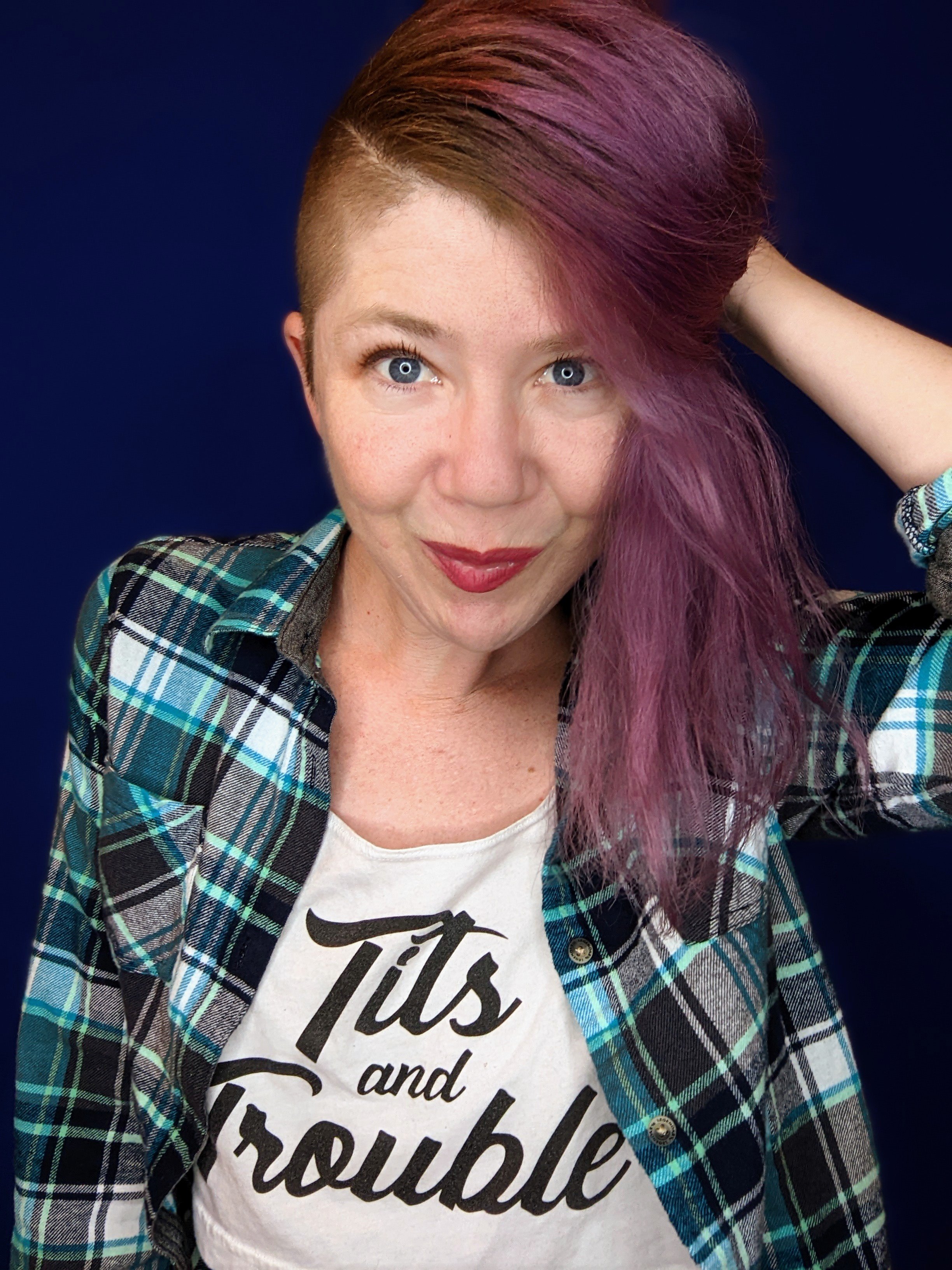 A person with short hair dyed purple and long hair with a purple streak, wearing a plaid shirt over a white t-shirt with black text, and looking at the camera with a slight smile against a dark blue background.