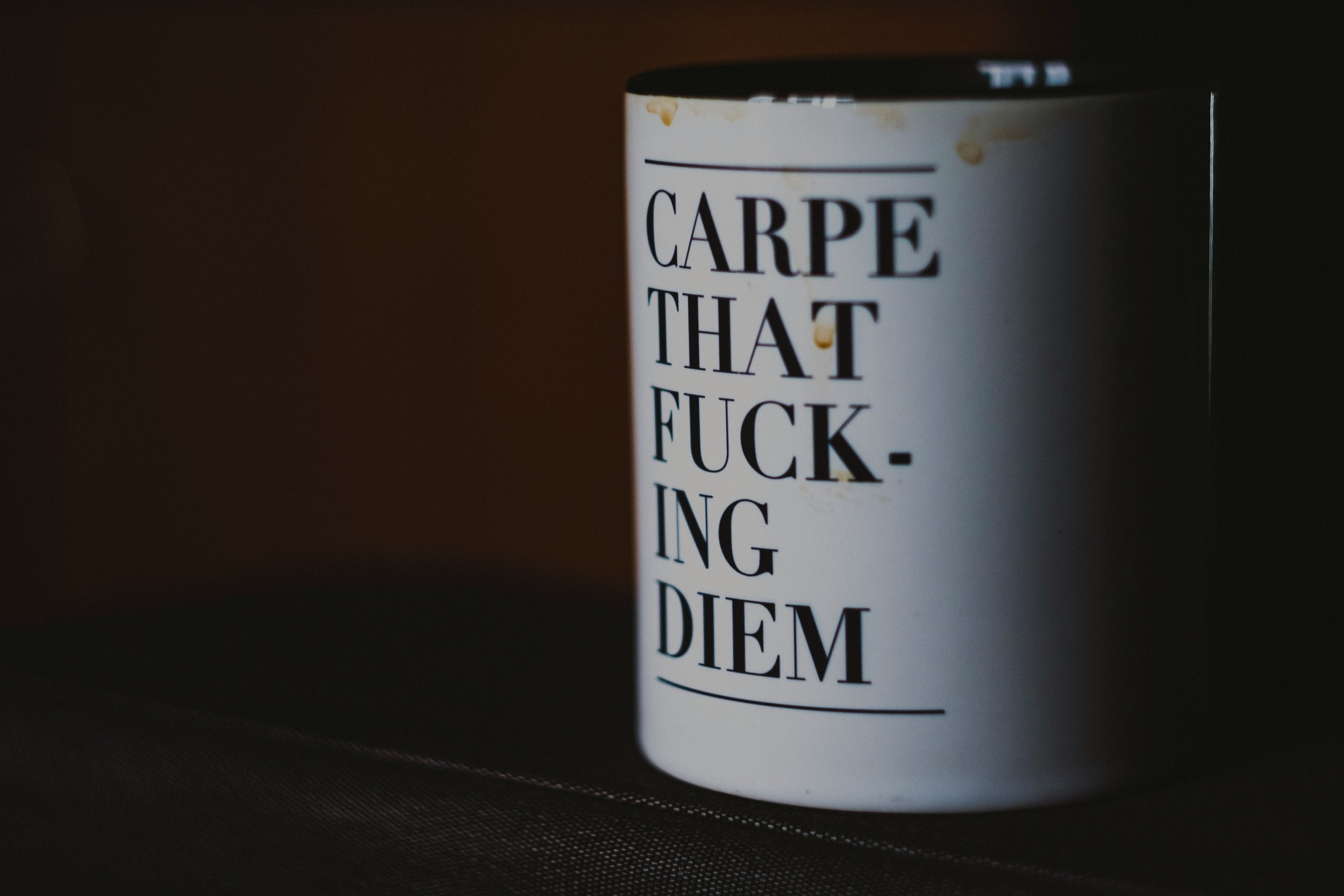 Close-up of a white mug with black text, partially visible, reading "Carpe that fucking diem," on a dark surface.
