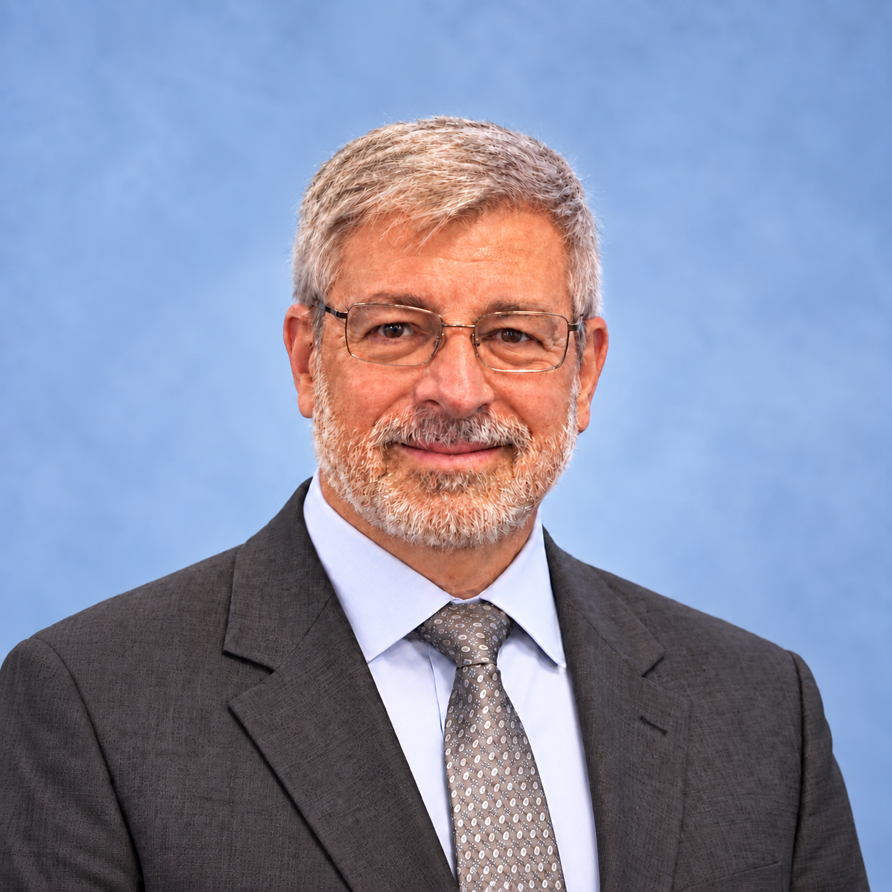 Headshot of a middle-aged man with gray hair, glasses, and a beard, wearing a dark suit, light blue shirt, and patterned tie, set against a blue background.
