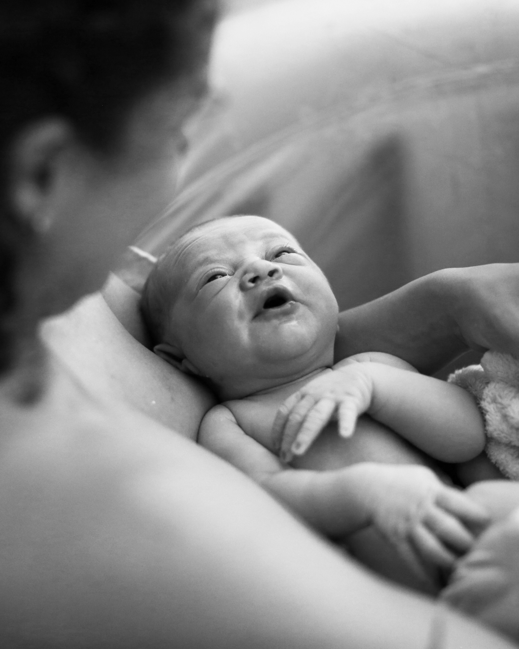 Un bébé regardant vers la caméra, tenu dans les bras d'une adulte, dans un environnement intérieur, en noir et blanc.