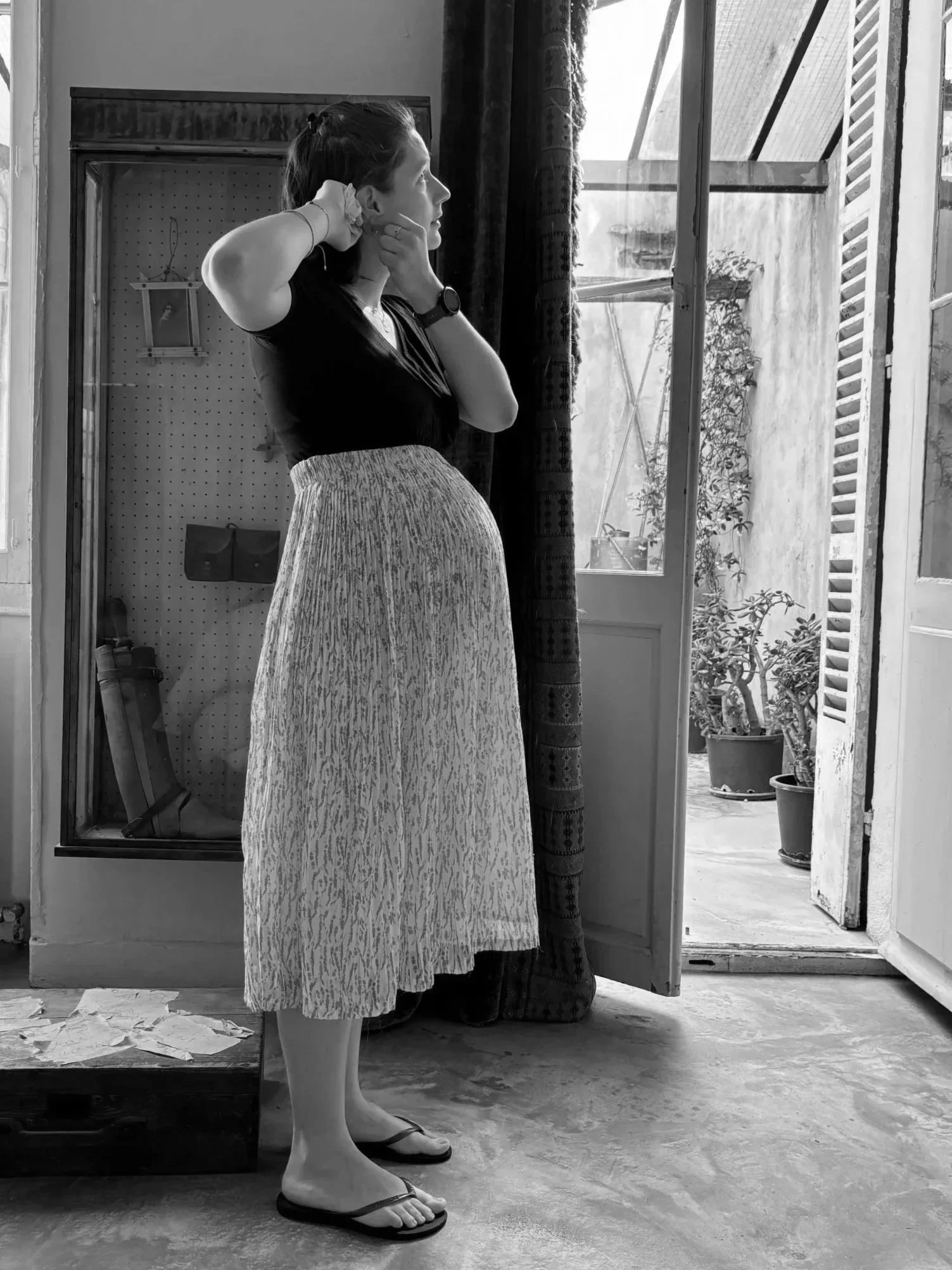 Une femme regarde par une fenêtre ouverte, debout dans une pièce avec un sol en ciment. Elle porte une jupe longue et un t-shirt noir, et elle ajuste ses écouteurs. La scène est en noir et blanc.