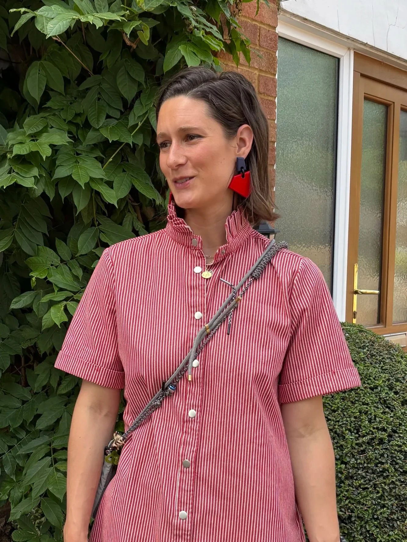 Femme avec des boucles d'oreilles rouges en forme de triangle, portant une robe à rayures rouges et blanches, avec une veste ou une sacoche en bandoulière grise, debout devant un mur de fleurs et une porte en bois.