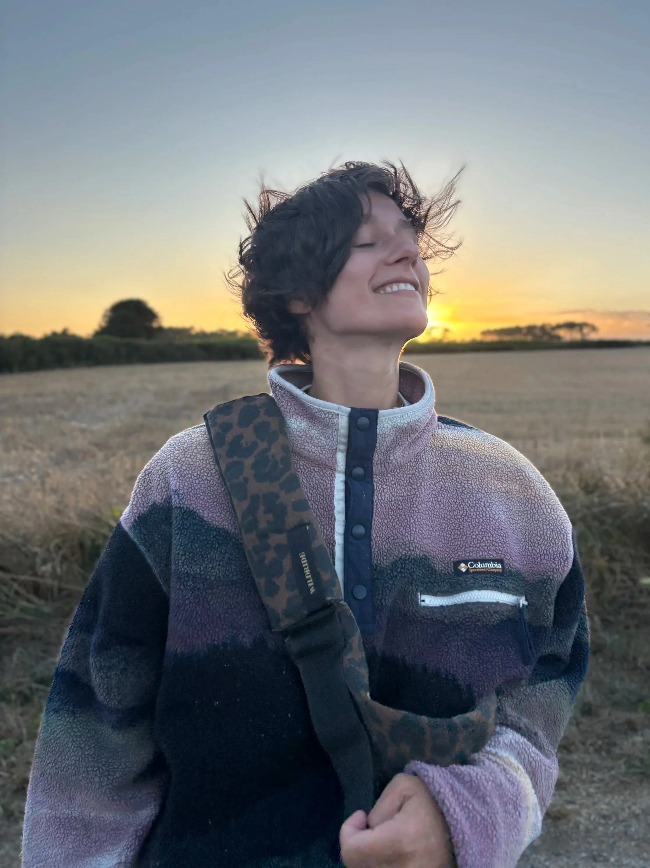 Une femme souriante avec les yeux fermés, portant une veste Columbia, dans un champ au coucher du soleil.