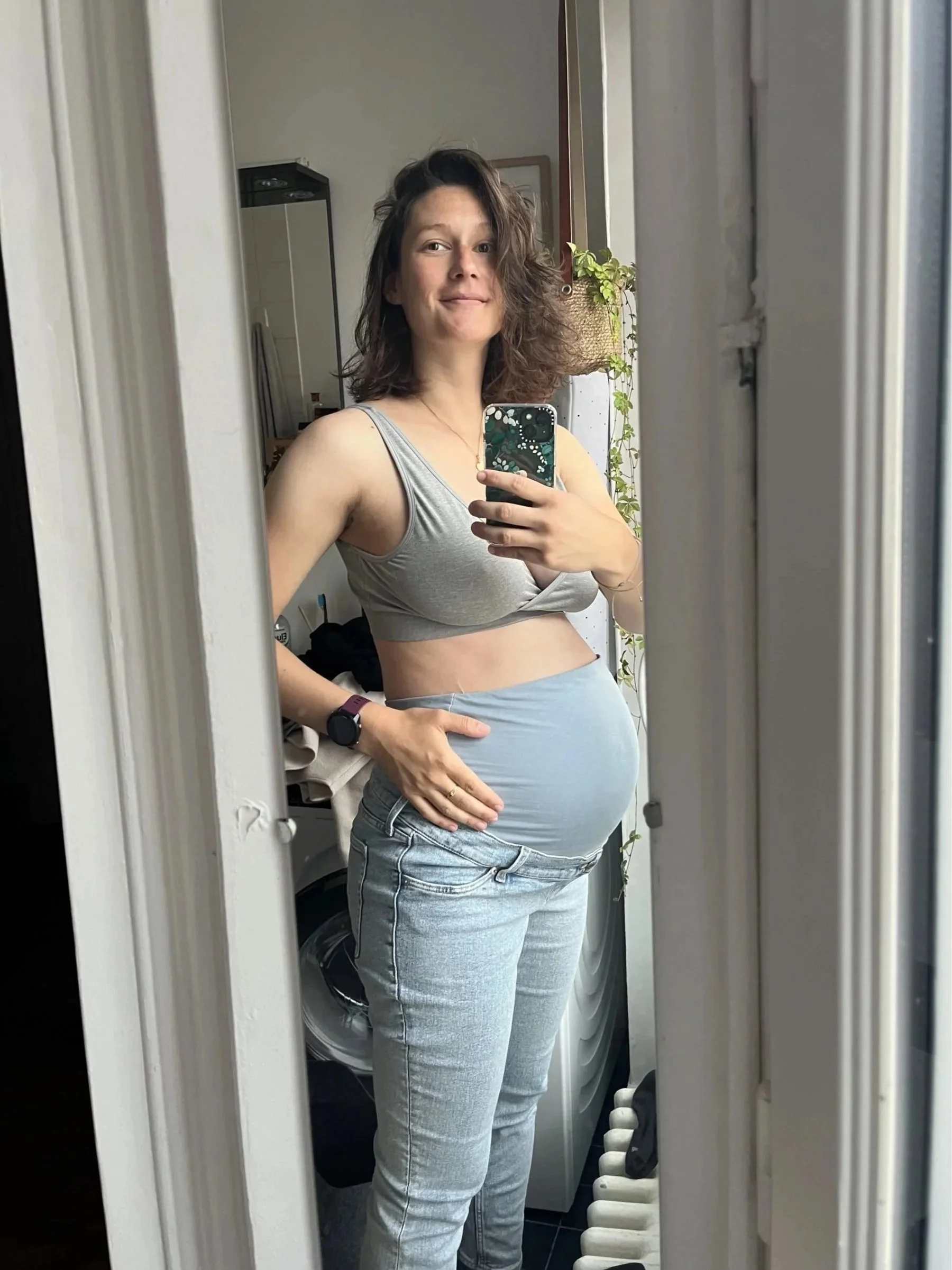 Femme prenant une selfie dans un miroir, montrant son ventre rond, dans une pièce avec plantes et meubles.