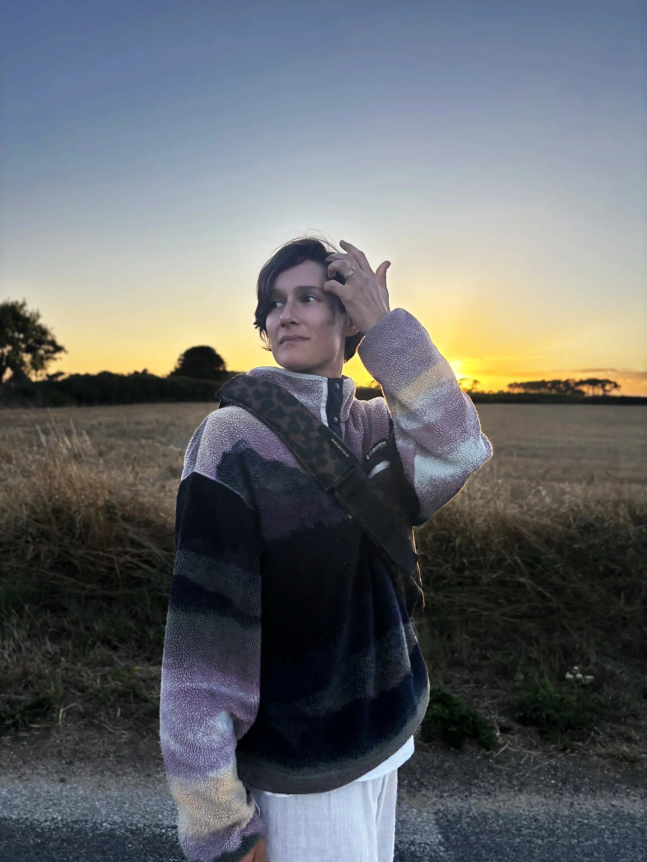 Une femme avec des cheveux courts et un pull coloré, posant à l'extérieur au coucher du soleil, avec un champ et des arbres en arrière-plan.