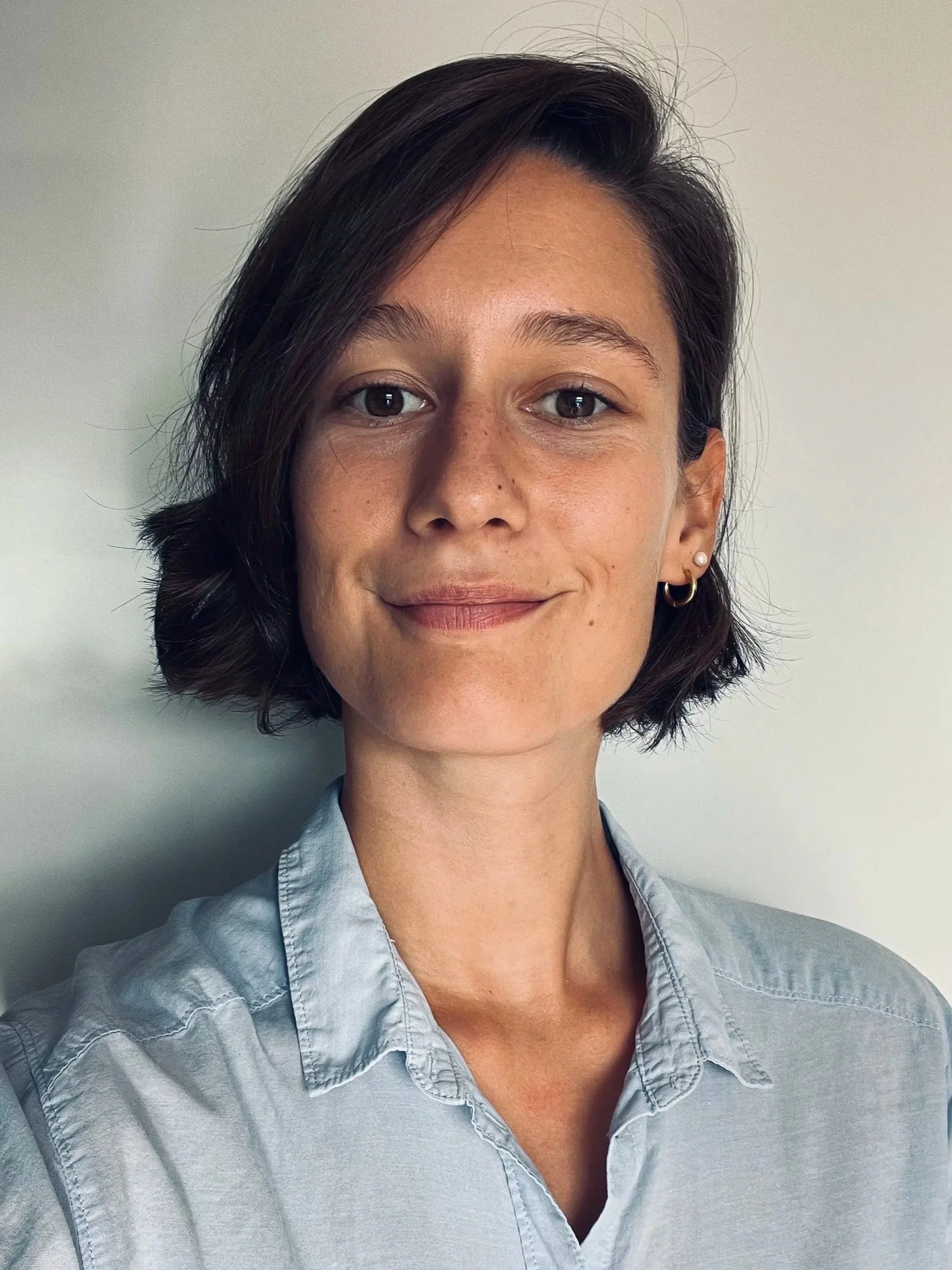 Jeune femme avec brune, cheveux courts et crépus, portant une chemise en denim claire, souriant légèrement, debout devant un mur blanc.
